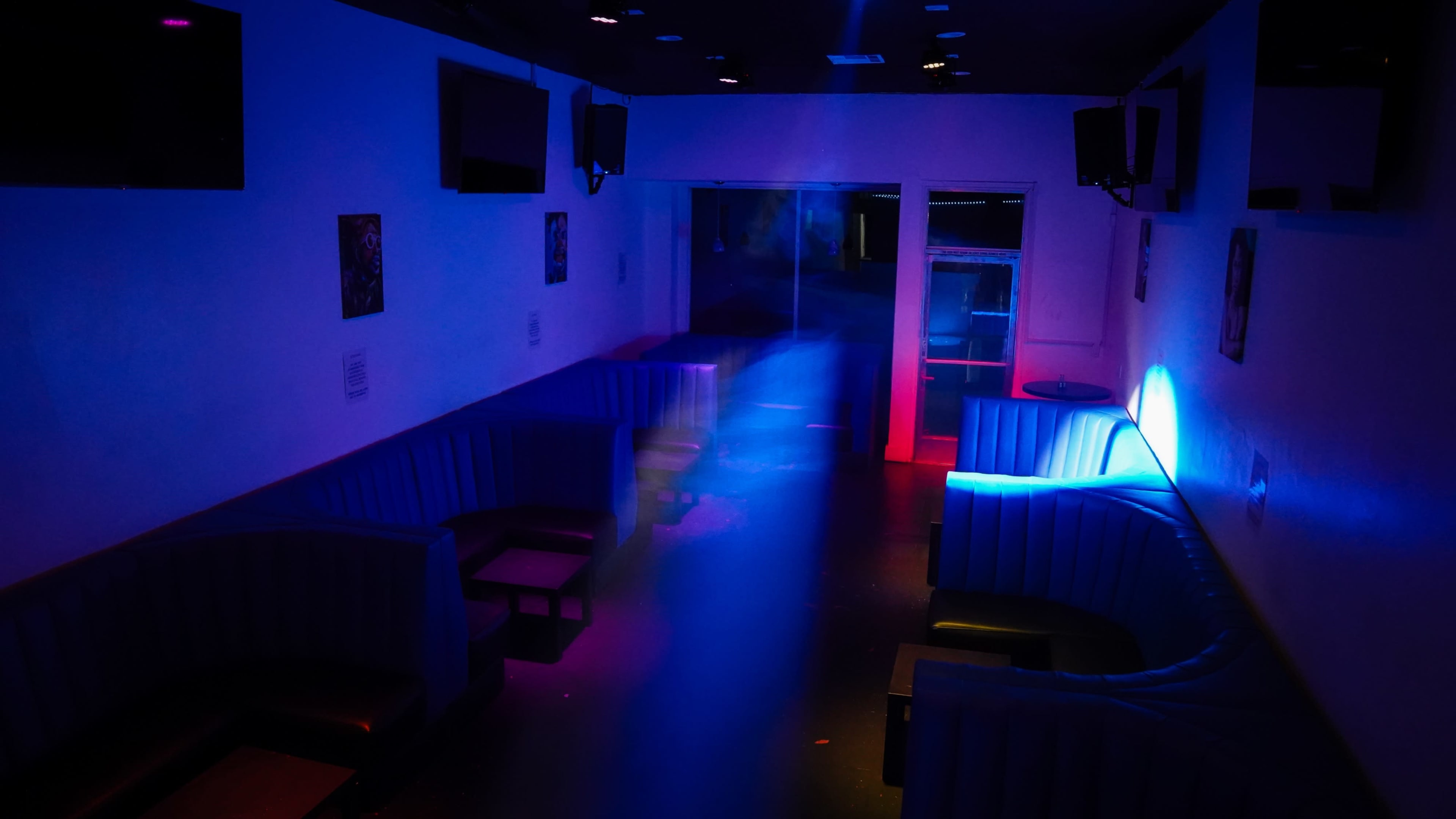 The image shows an empty lounge space with blue and red lighting, featuring circular booths against the walls.