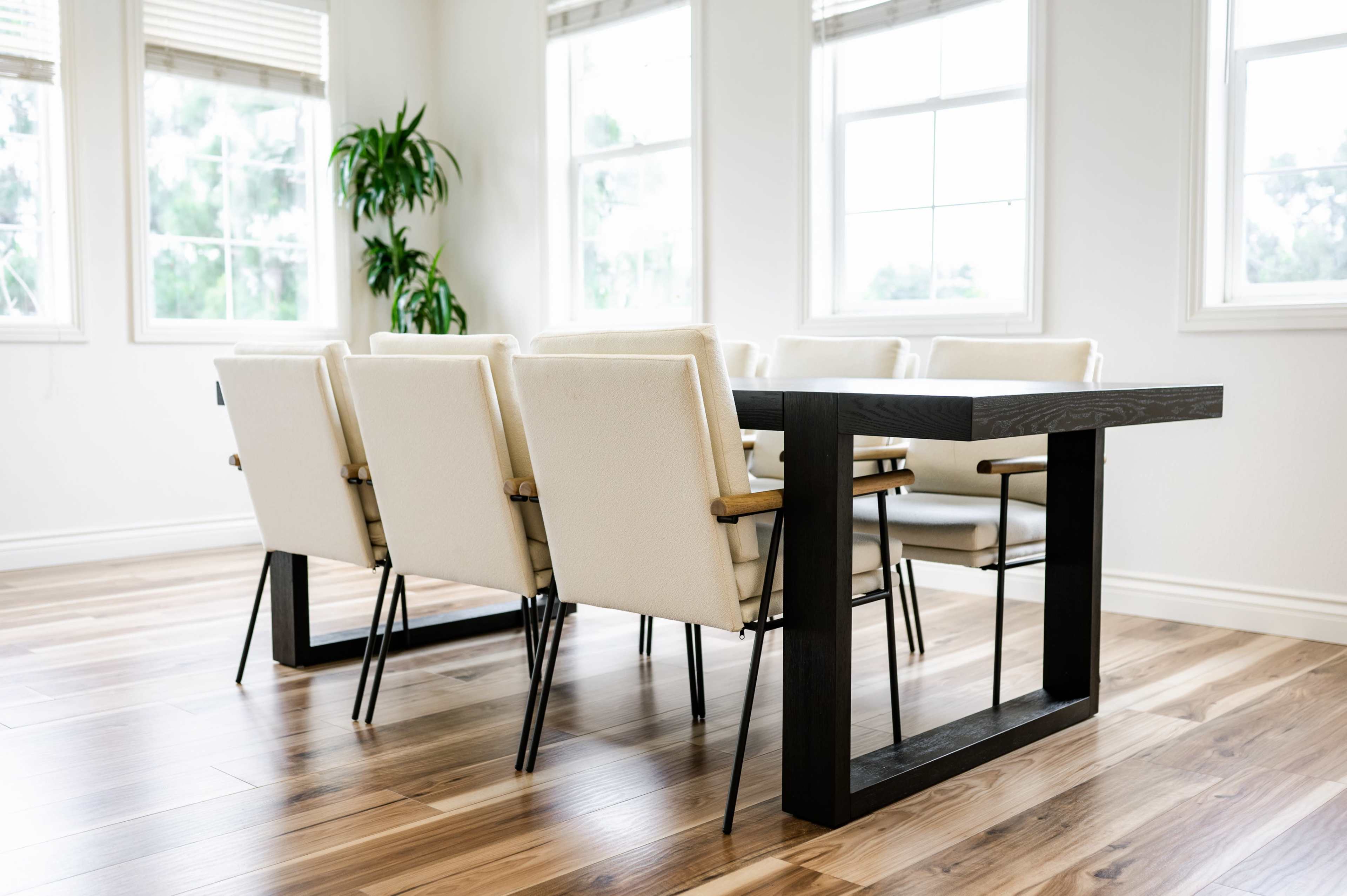 A modern dining room features a black rectangular table surrounded by several white upholstered chairs, with large windows and a green plant in the background.