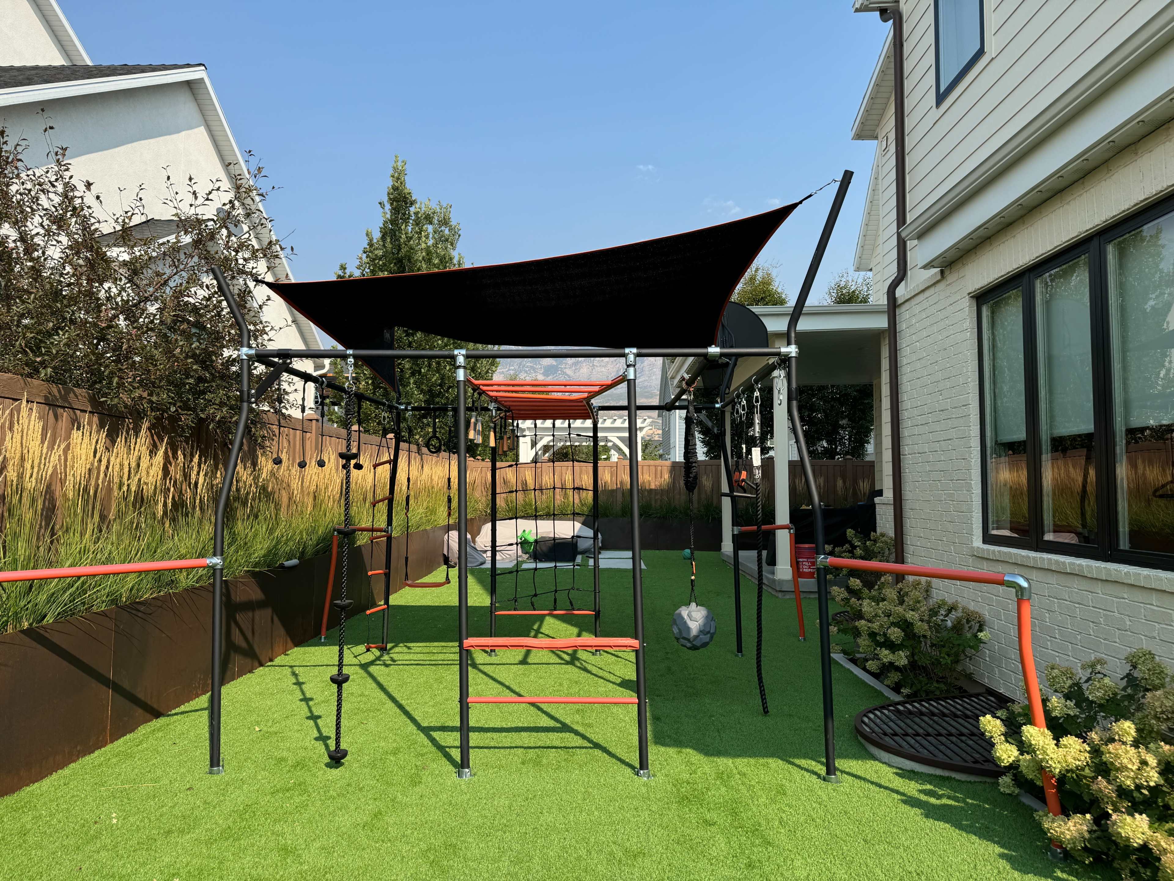The image shows a playground structure with various climbing apparatuses set on green artificial grass in a backyard.