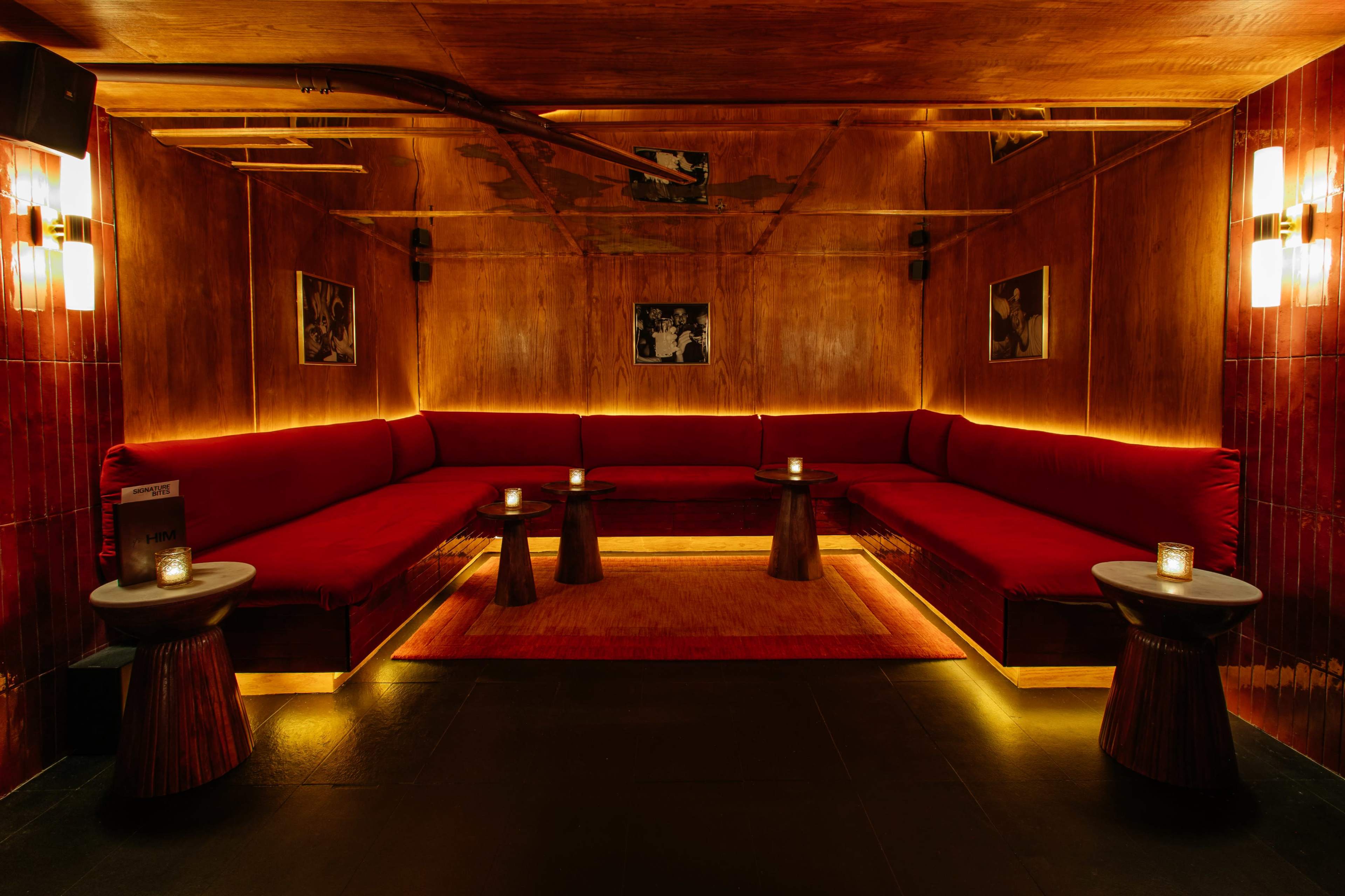 The image shows a dimly lit lounge area featuring red velvet seating against wooden walls, with low tables and flickering candles on them.