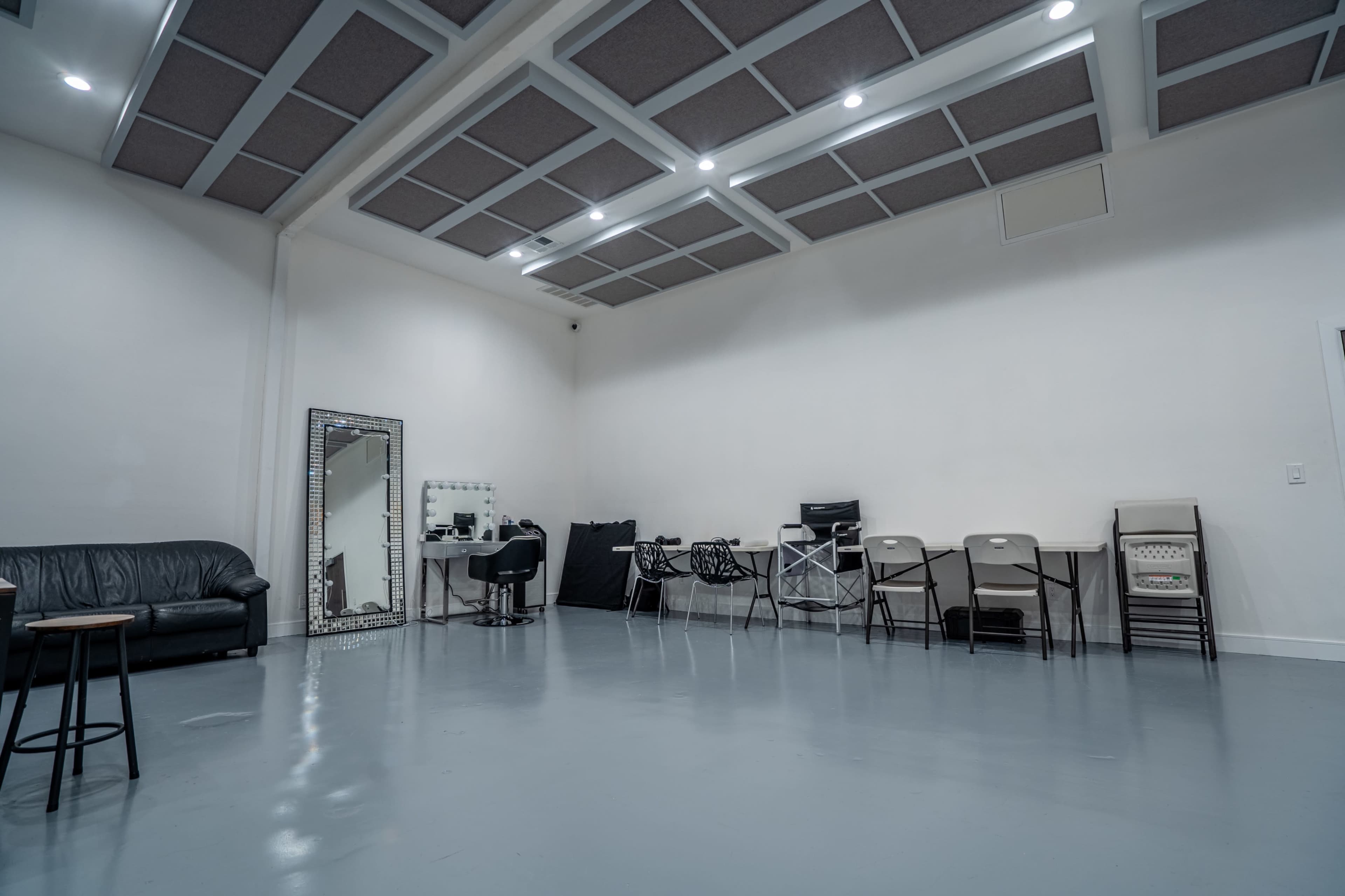 The image shows a spacious, empty room featuring a black leather couch, a mirror, and several folding chairs along the wall.