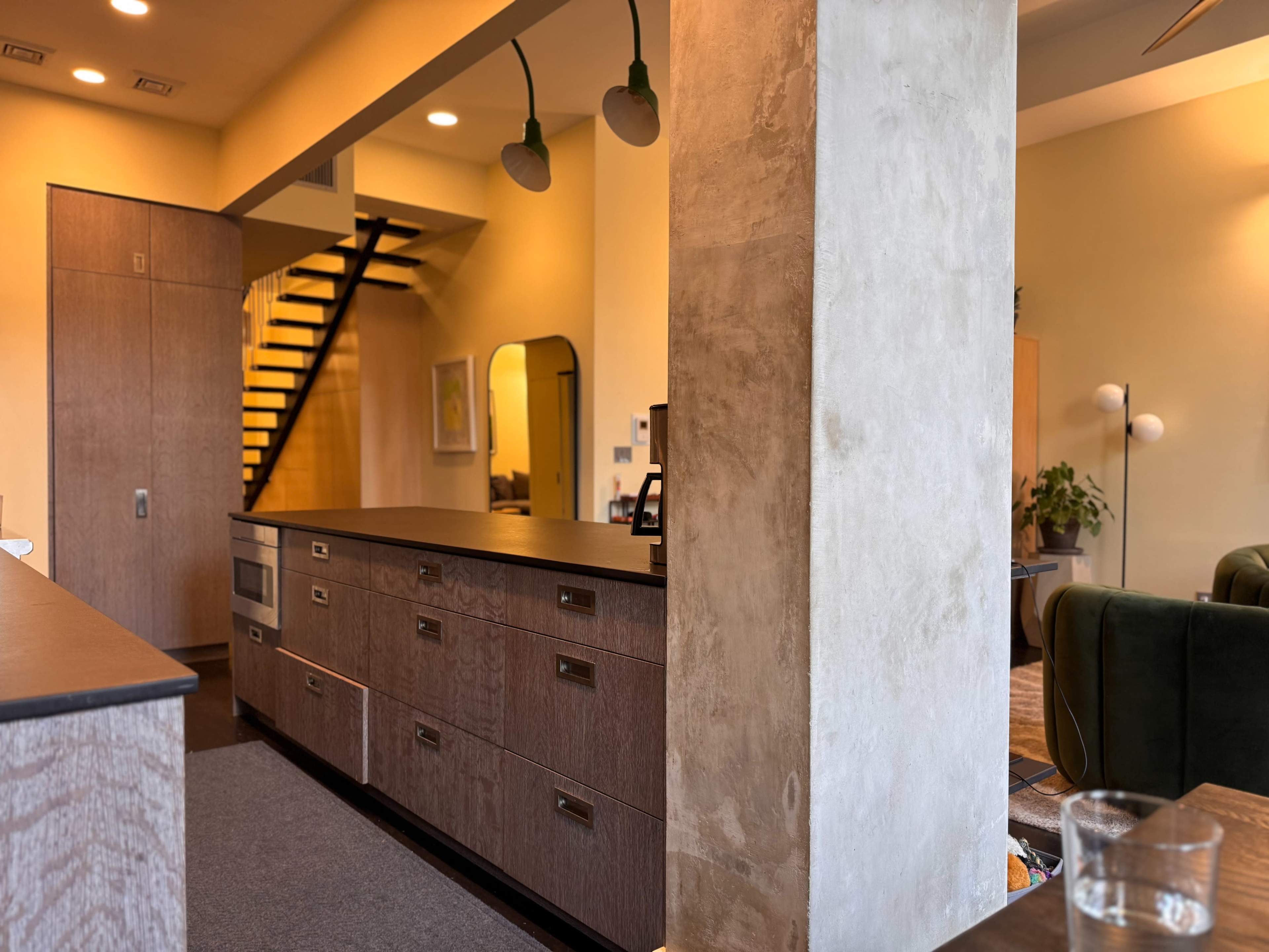 The image shows a modern interior space with a wooden kitchen island, a staircase in the background, and a mixture of textures on the walls.