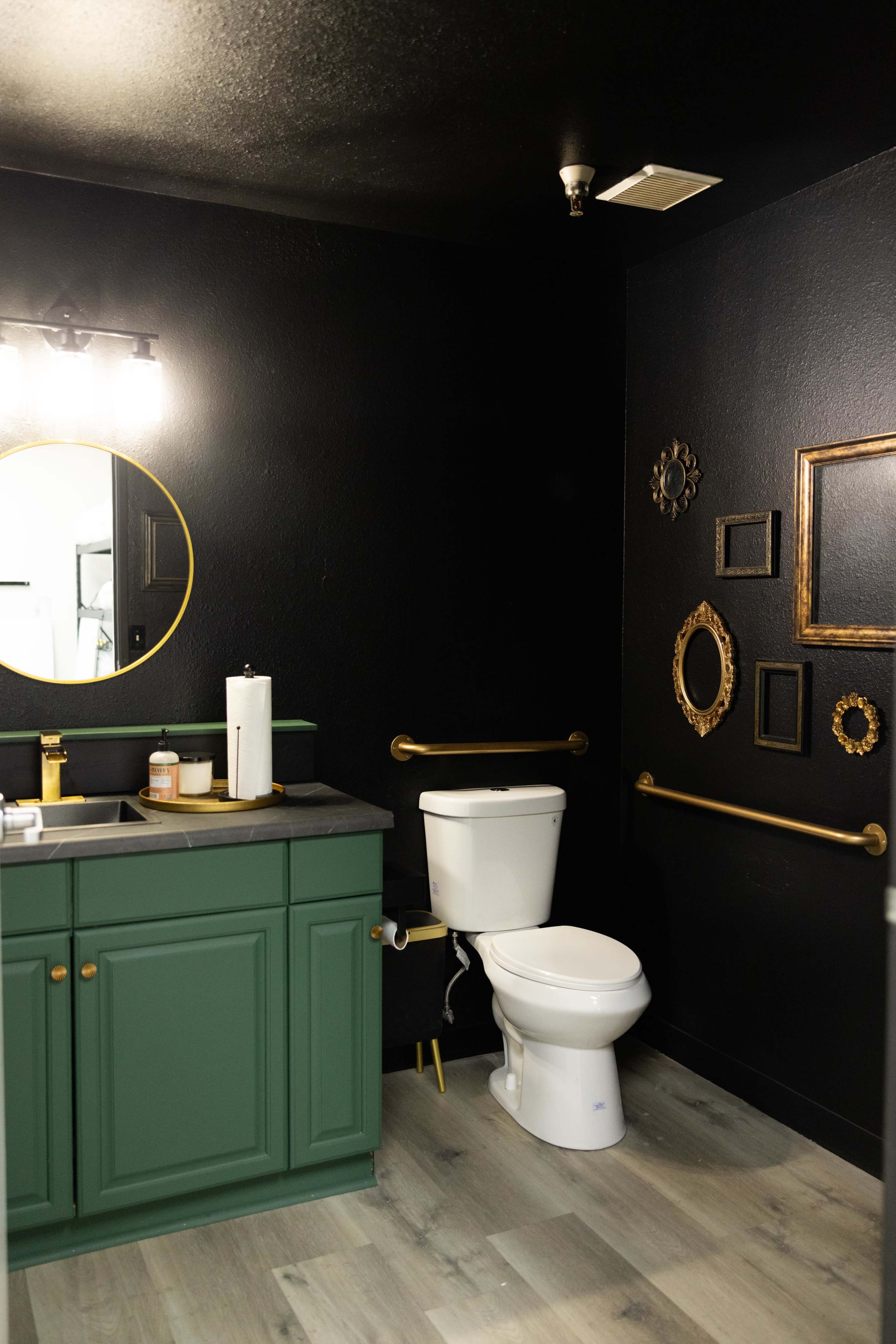 The image shows a modern bathroom featuring a green vanity, a white toilet, gold handrails, and a black wall adorned with decorative frames.