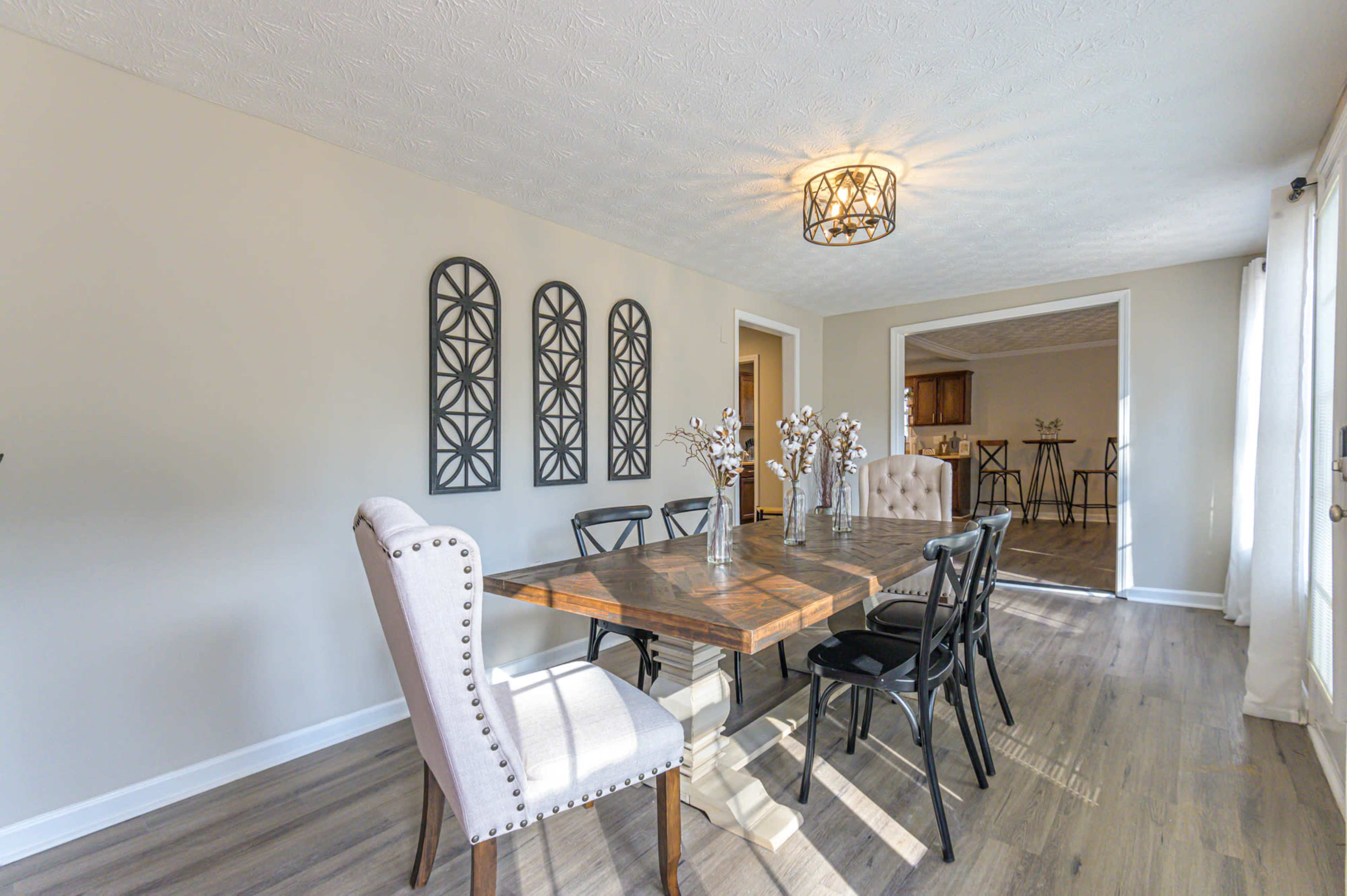 A dining room features a wooden table surrounded by black and beige chairs, with decorative wall panels and a chandelier above.