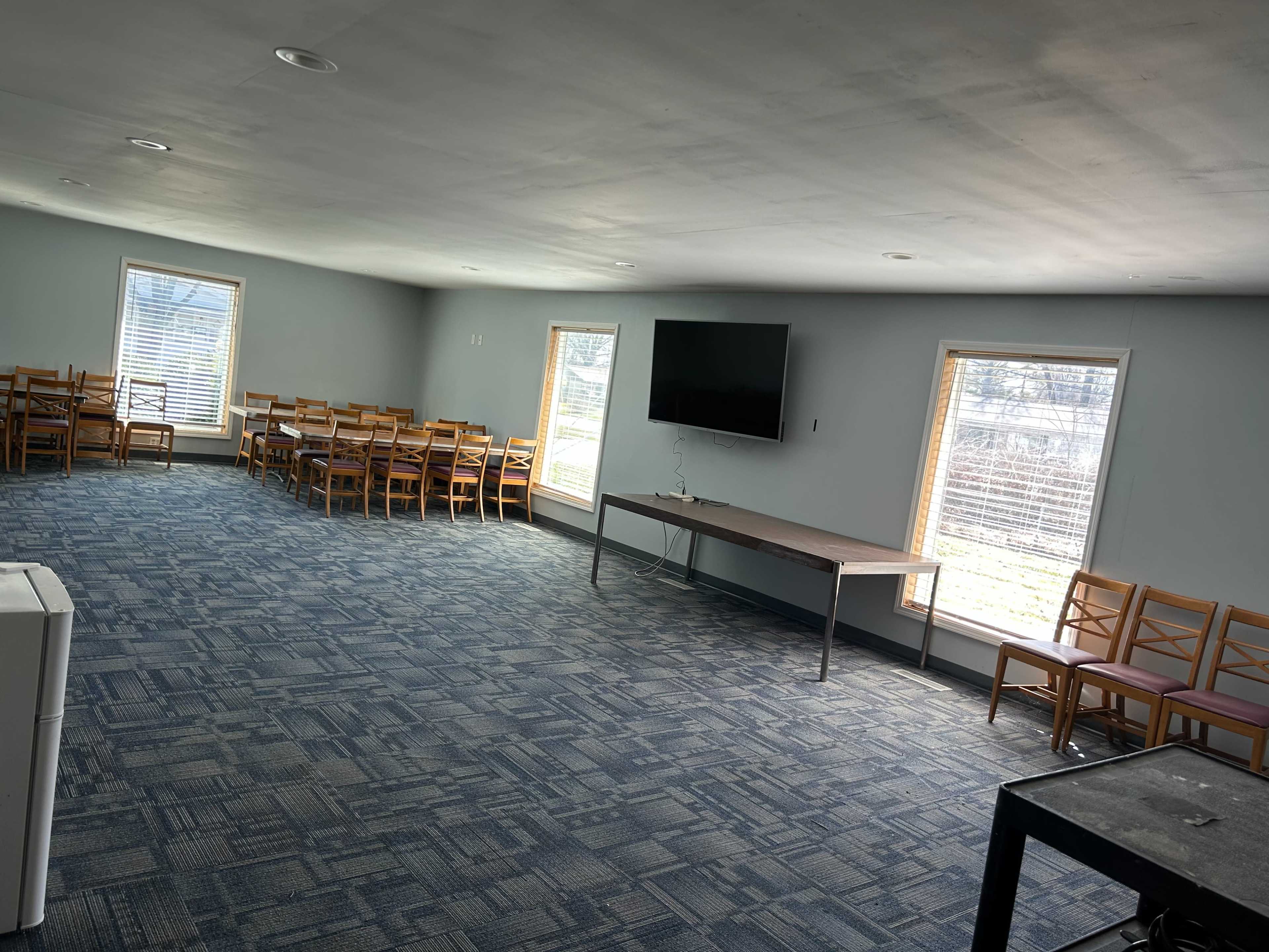 The image shows a spacious, well-lit room with rows of wooden chairs, a large table, and a television mounted on the wall.