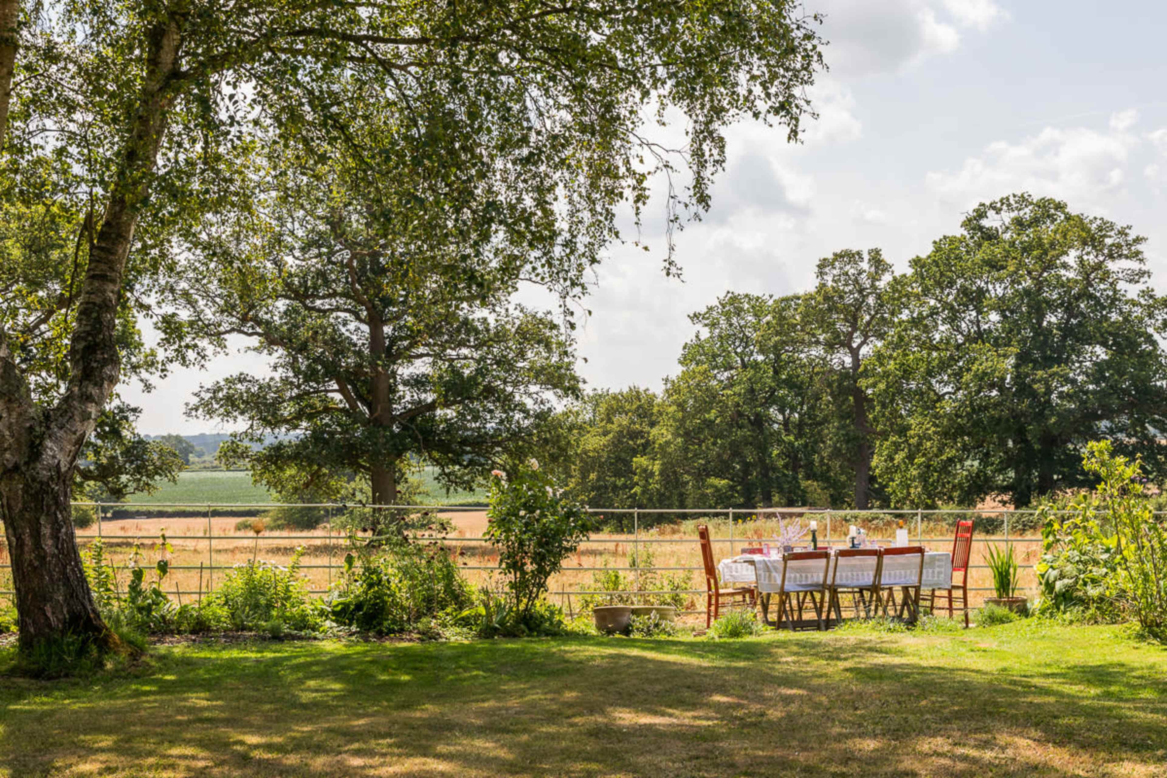 A table set for a meal is positioned under trees, overlooking a grassy area with fields in the background.