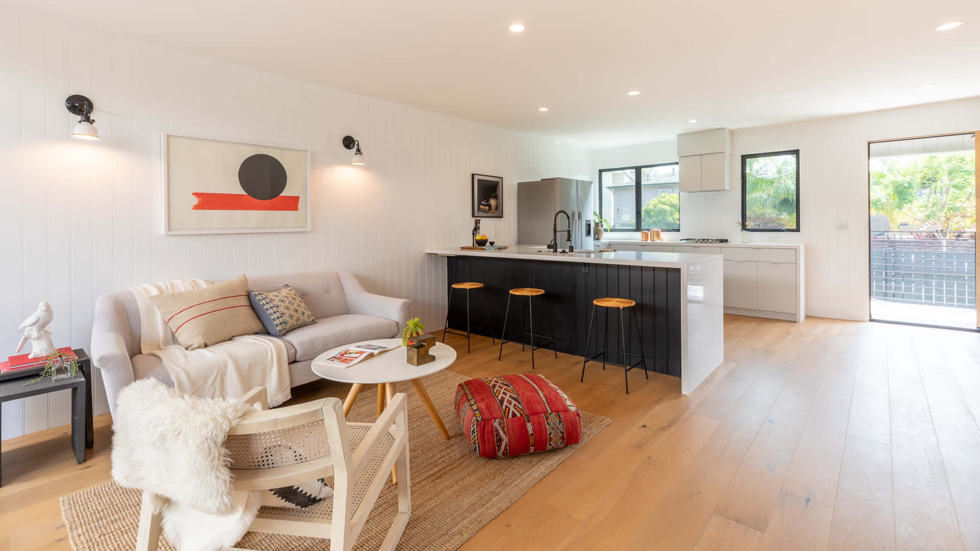 The image displays a modern, open-concept living area with a gray sofa, a round coffee table, and a kitchen in the background featuring stainless steel appliances and bar stools.