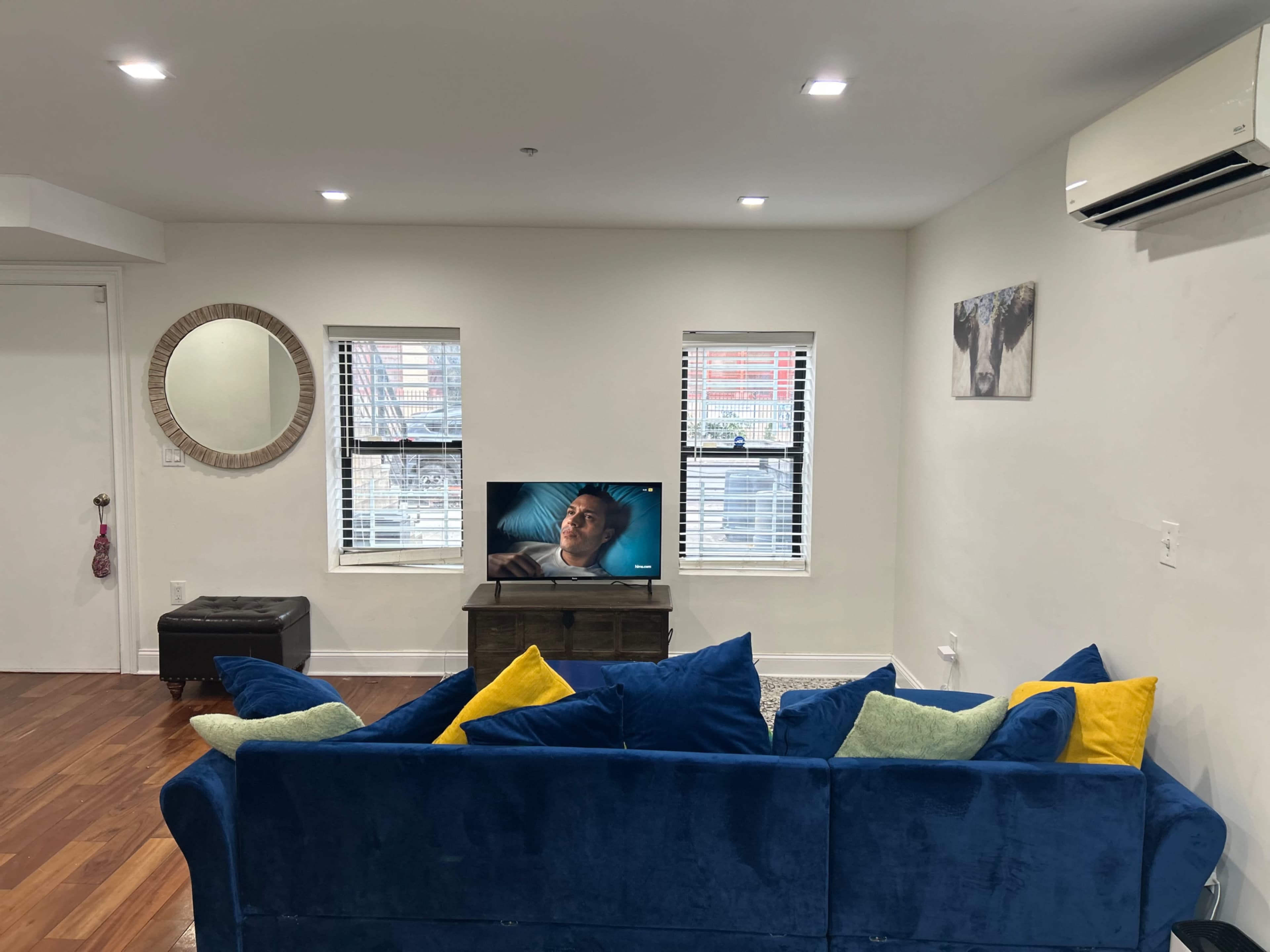 The living room features a blue sofa adorned with colorful pillows, a wooden television stand with a television facing it, and two windows allowing natural light to enter the space.