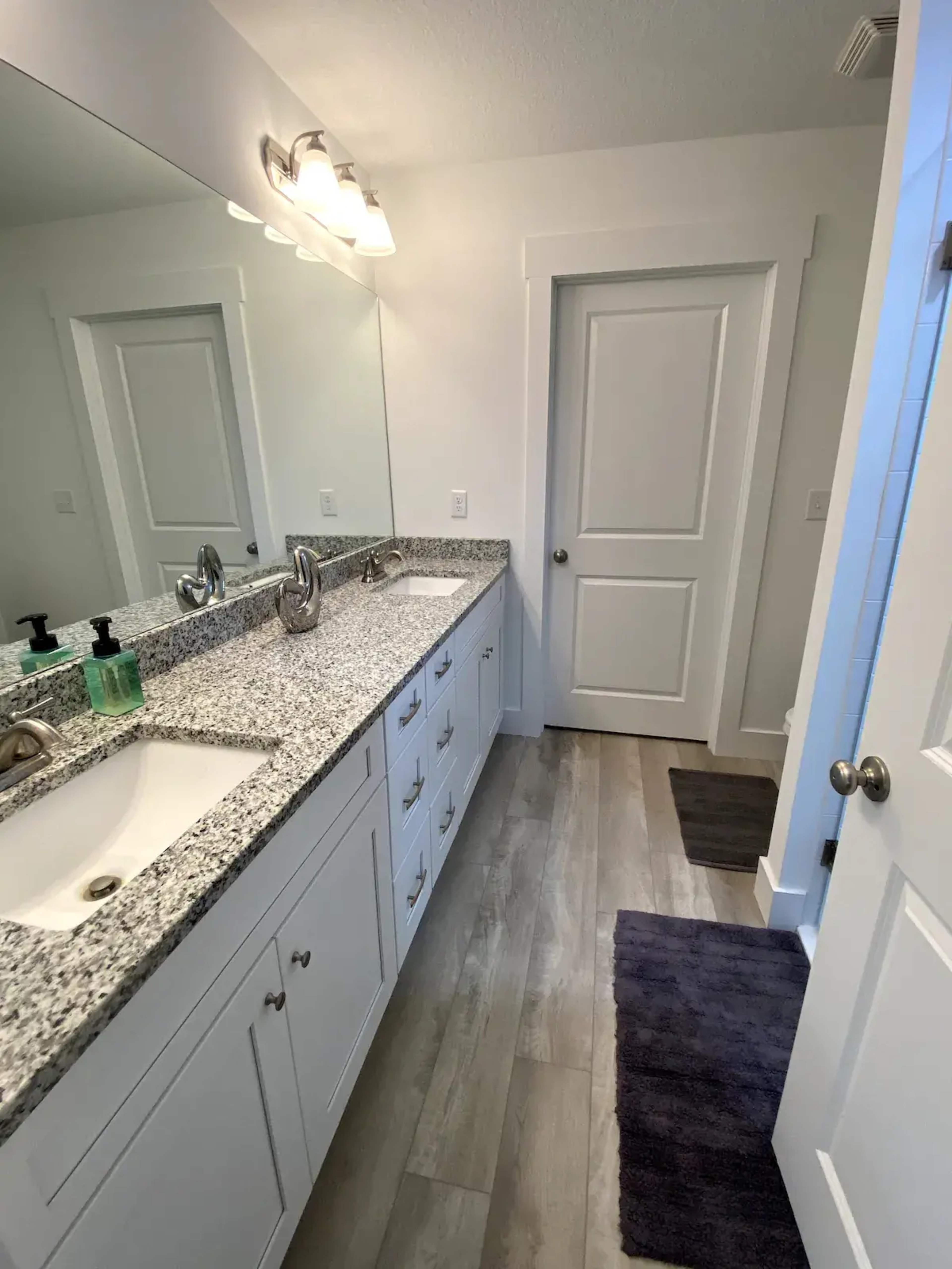 A clean bathroom featuring a double vanity with a granite countertop, two sinks, and a wall-mounted mirror, alongside a door leading to another room.