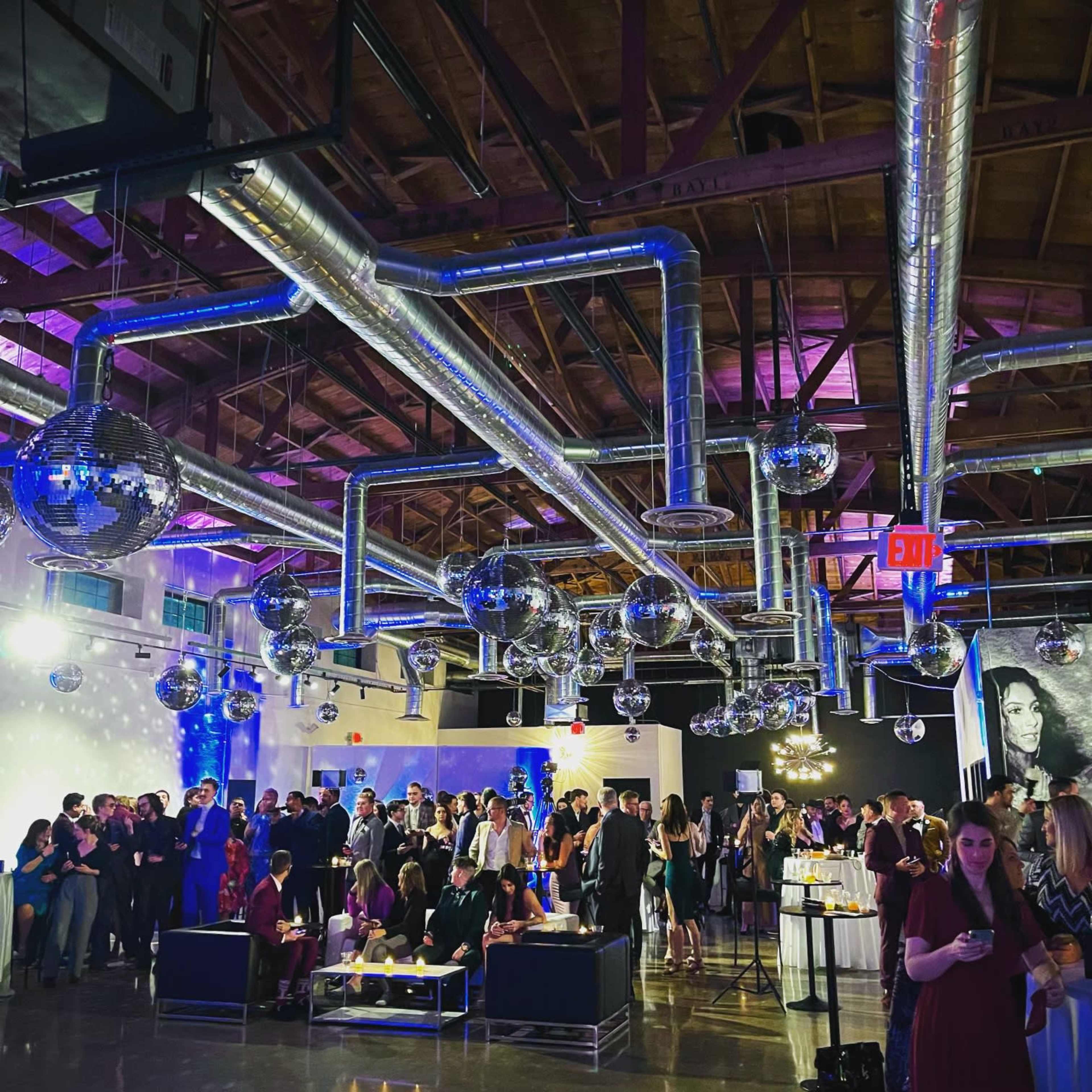 A large gathering takes place in a stylish venue featuring disco balls, exposed ductwork, and people mingling around tables.