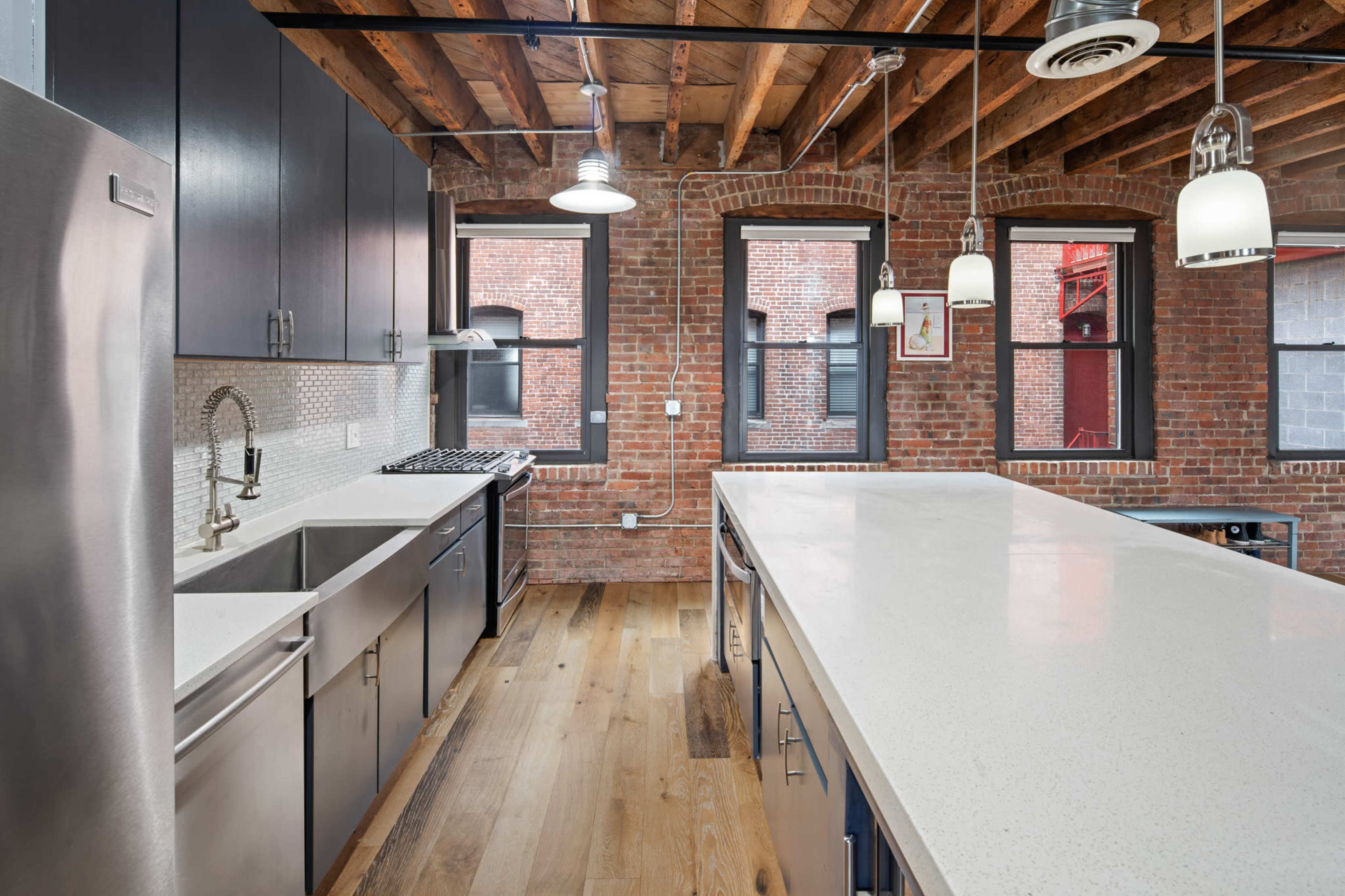 A modern kitchen featuring stainless steel appliances, a large island with a white countertop, and exposed brick walls with wooden beams overhead.