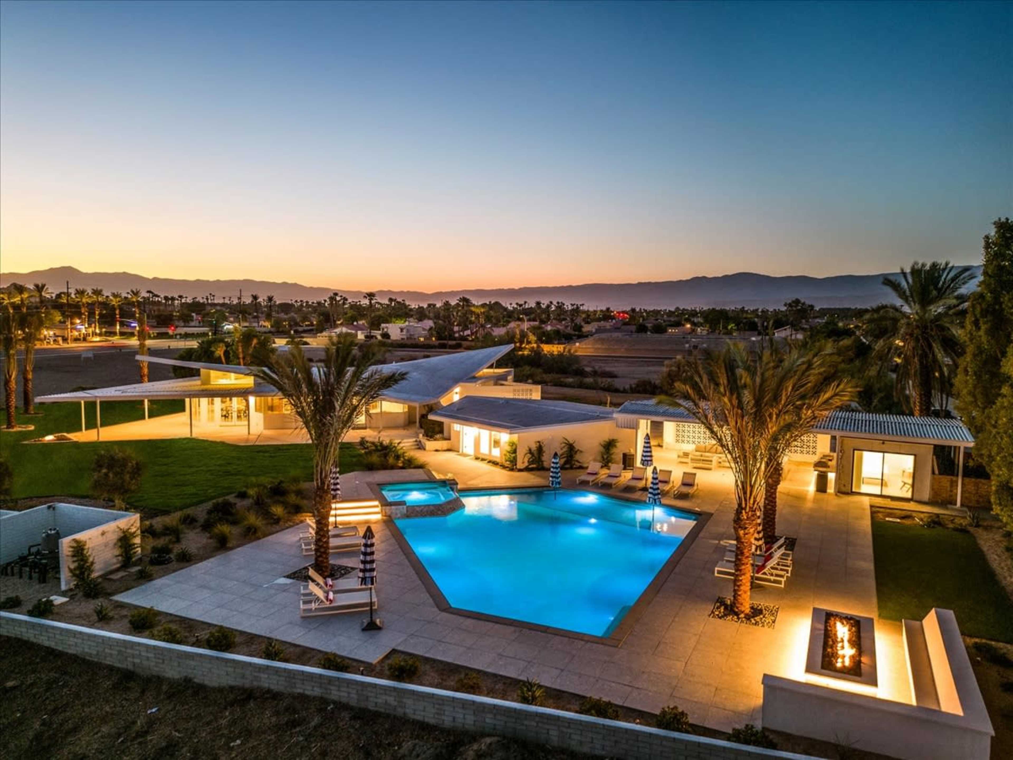 A modern house with a swimming pool, palm trees, and a fire pit is illuminated under a twilight sky.
