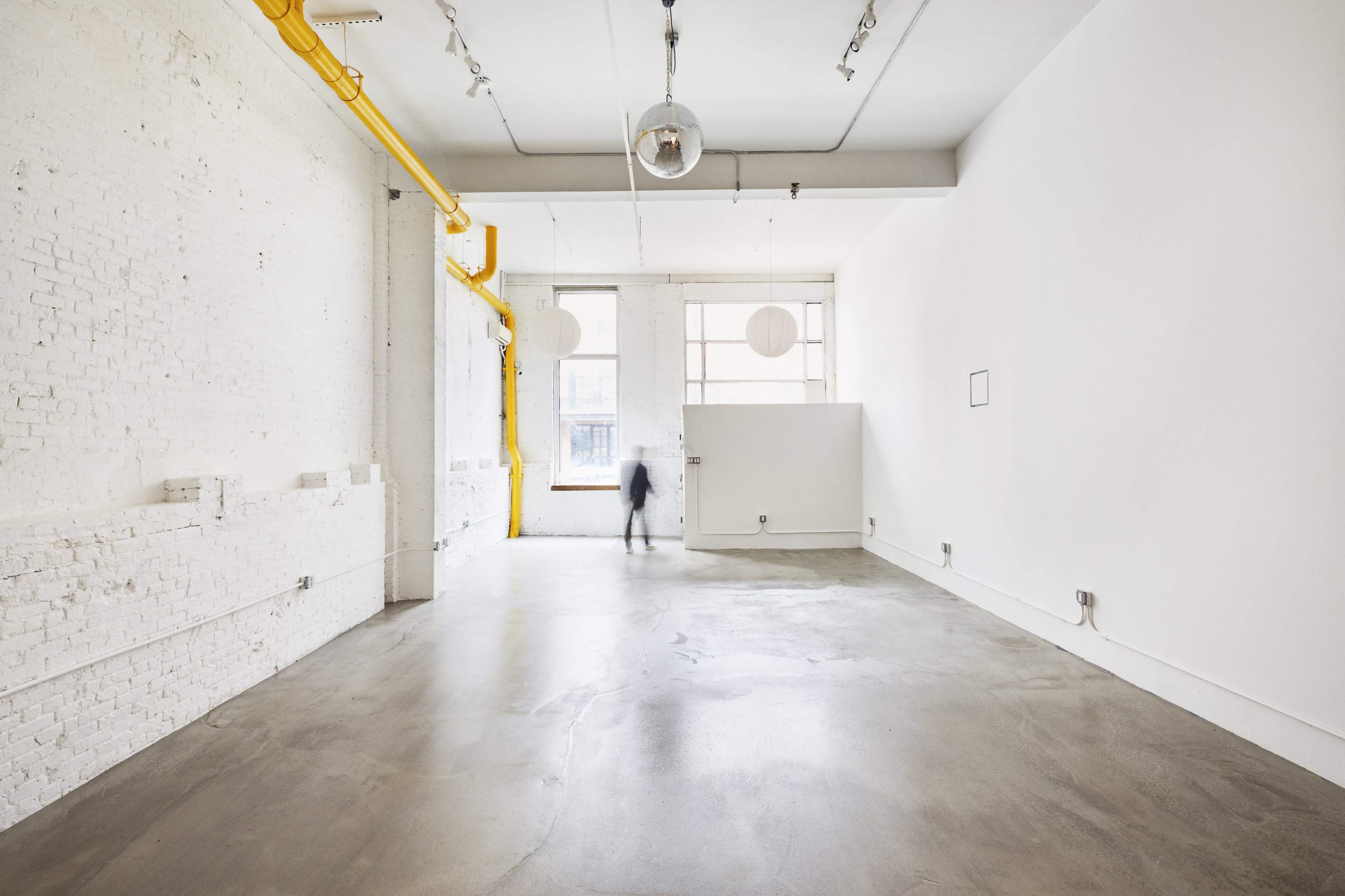 A spacious, empty room with white brick walls, a concrete floor, and large windows allowing natural light to enter.