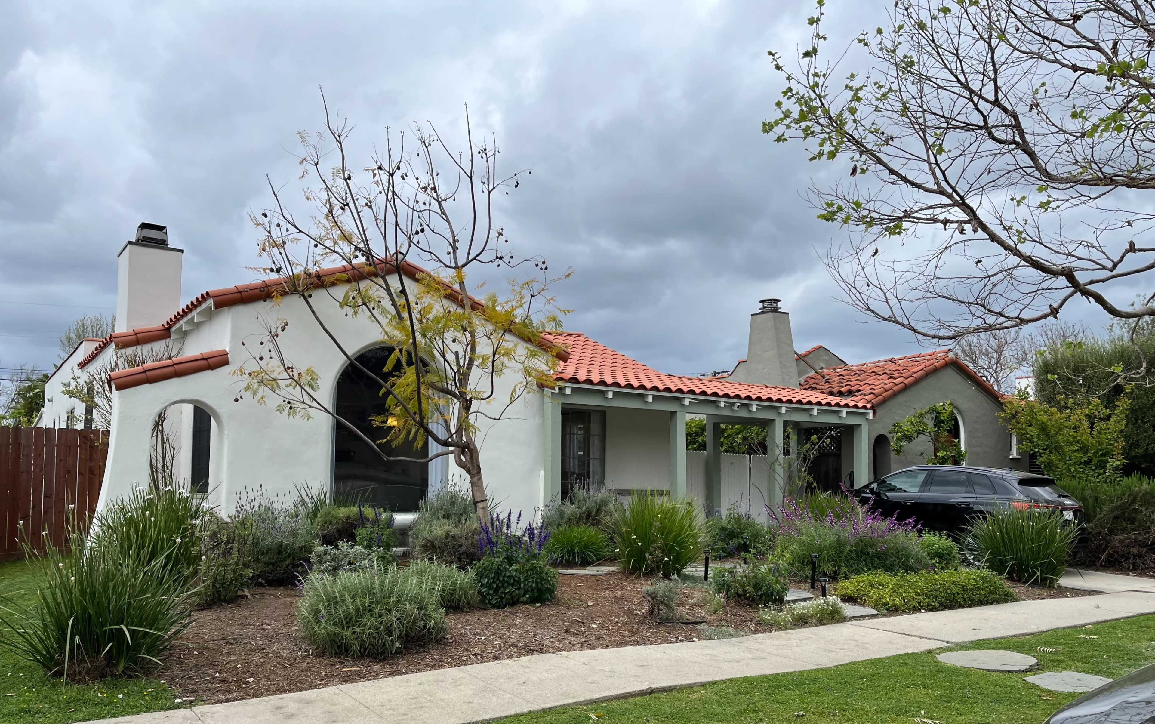 A white stucco house with a red-tiled roof is surrounded by landscaped gardens and a parked car in the driveway.