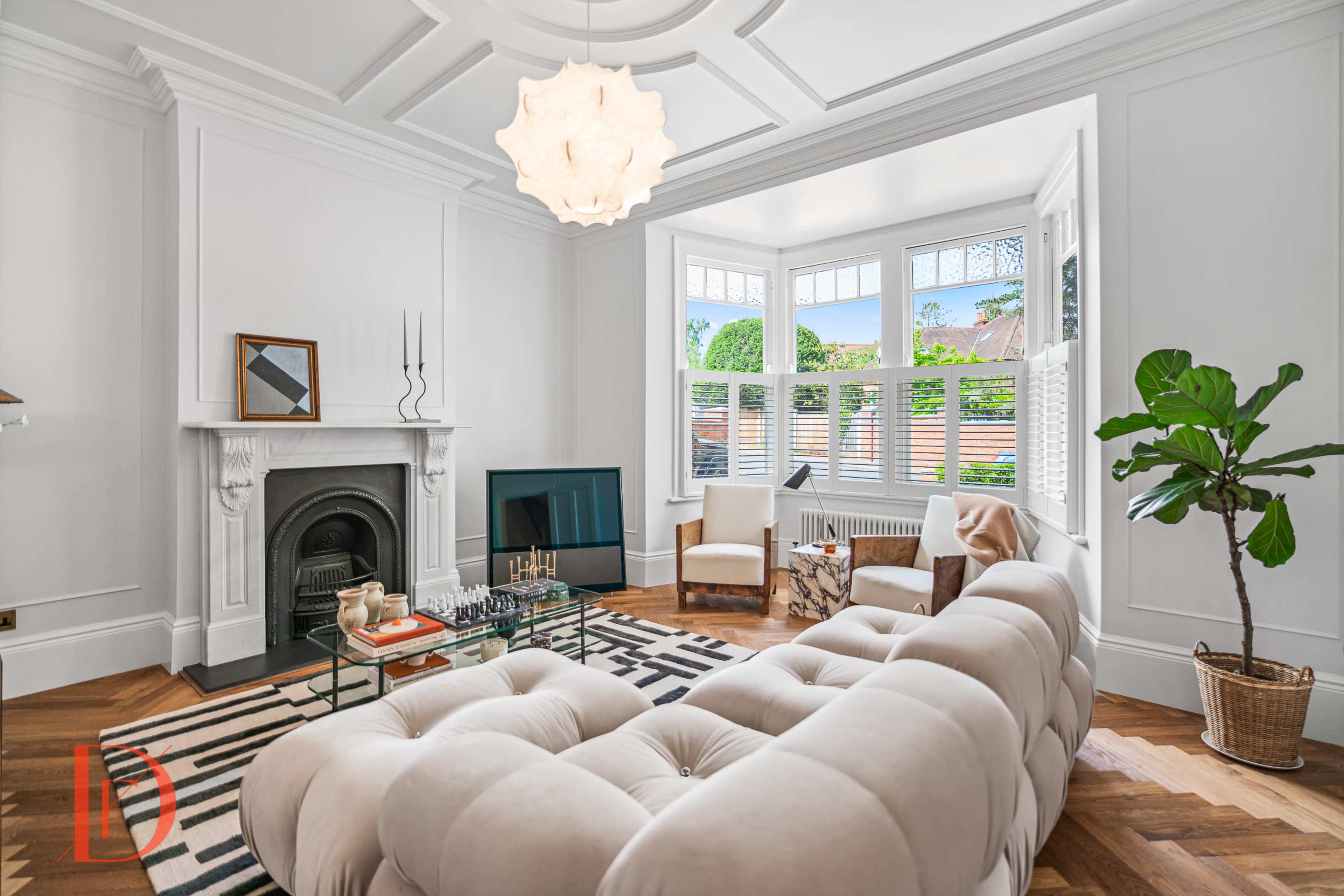 A well-lit living room featuring a tufted beige sofa, a glass coffee table, and large windows with shutters overlooking a garden.