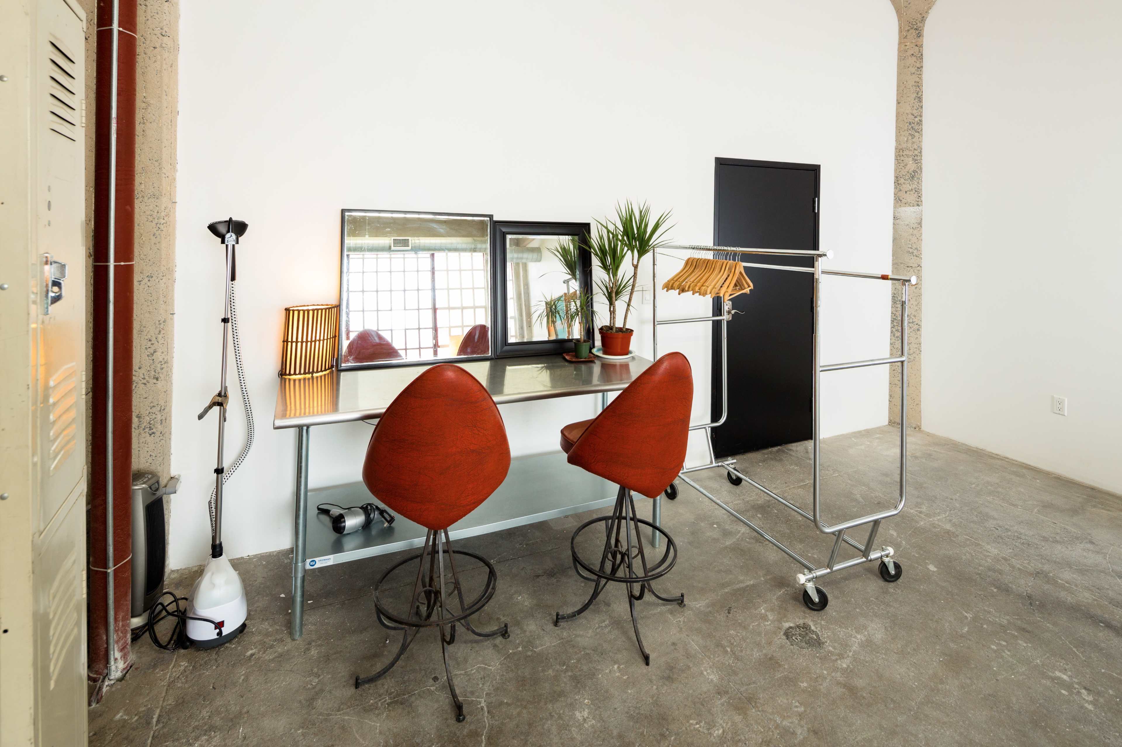 A minimalistic workspace features a glass desk with two red chairs, a wall-mounted mirror, a potted plant, and a clothing rack on wheels.