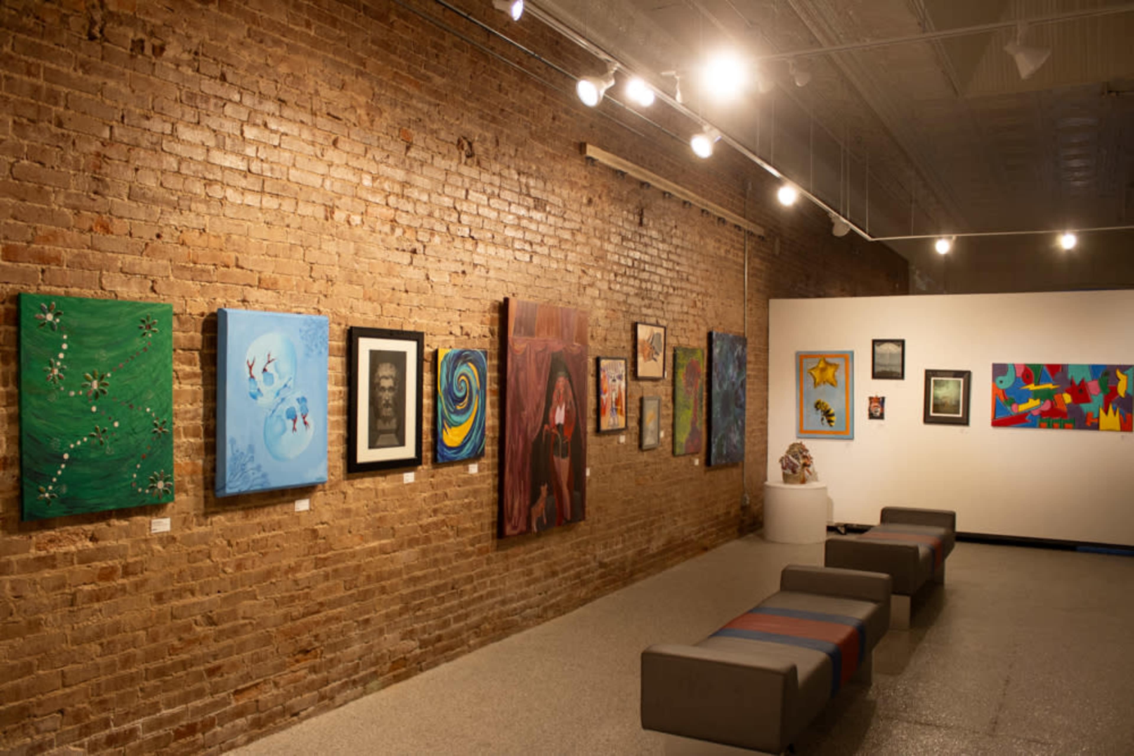 The image shows an art gallery with a brick wall, featuring various paintings hung on the wall and a row of benches in the foreground.