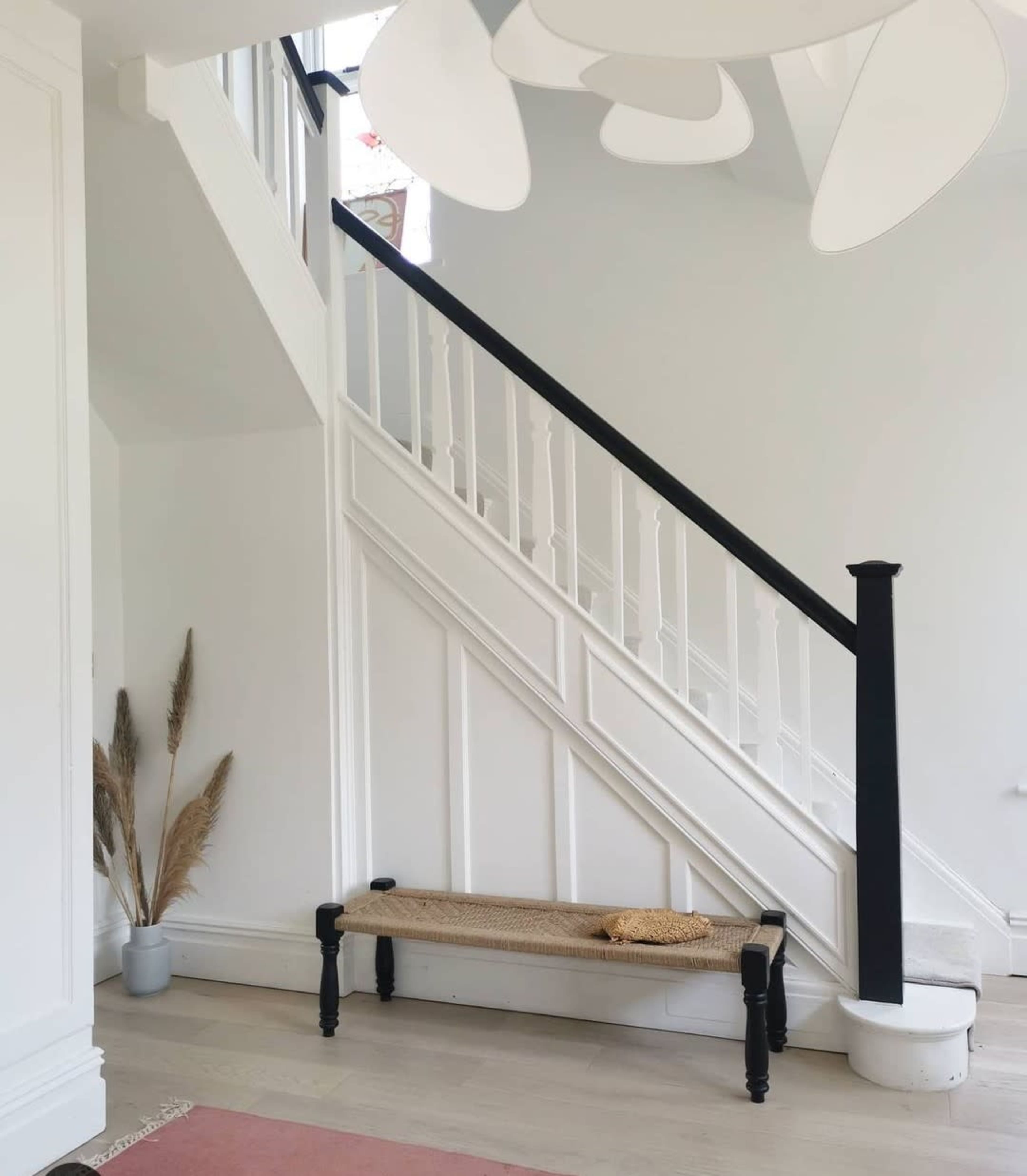 The image shows a bright entryway featuring a staircase with black railing, a woven bench against the wall, and decorative plants in the corner.