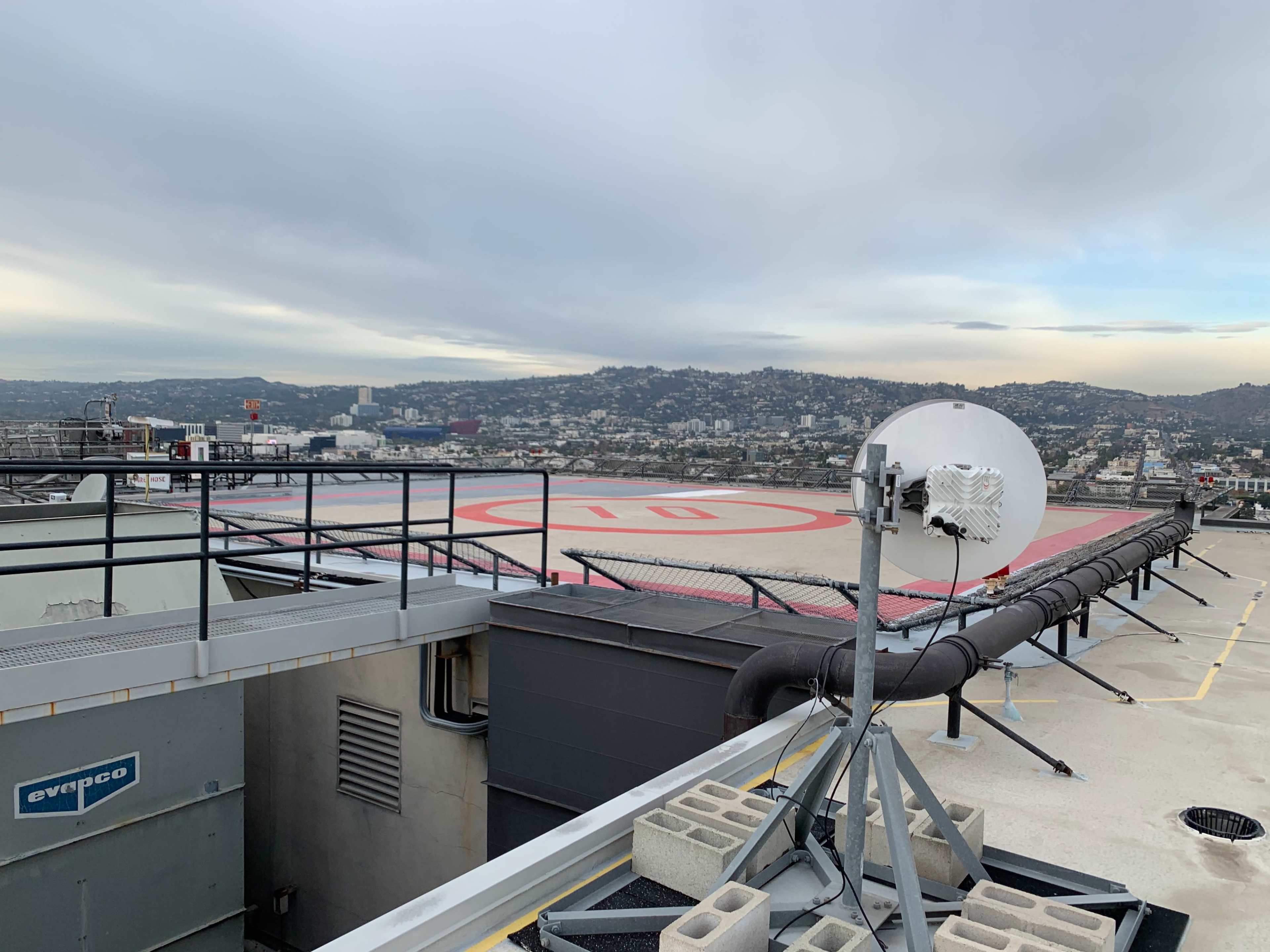 A rooftop helipad overlooks a cityscape with hills in the background and a satellite dish mounted nearby.