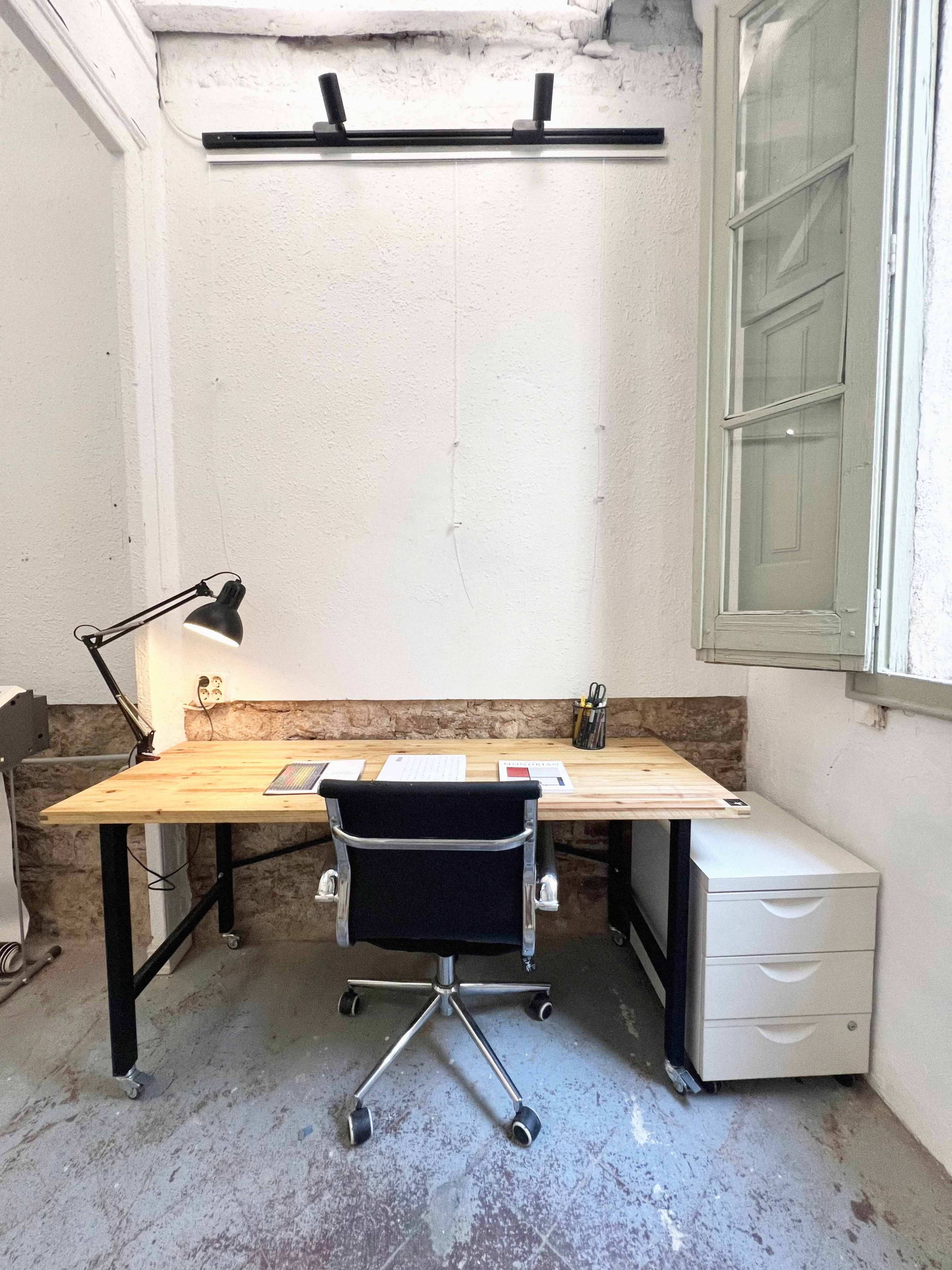 The image shows a minimalist workspace featuring a wooden desk with a swivel chair, a desk lamp, stationery, and a filing cabinet, all set against a white wall and a window.