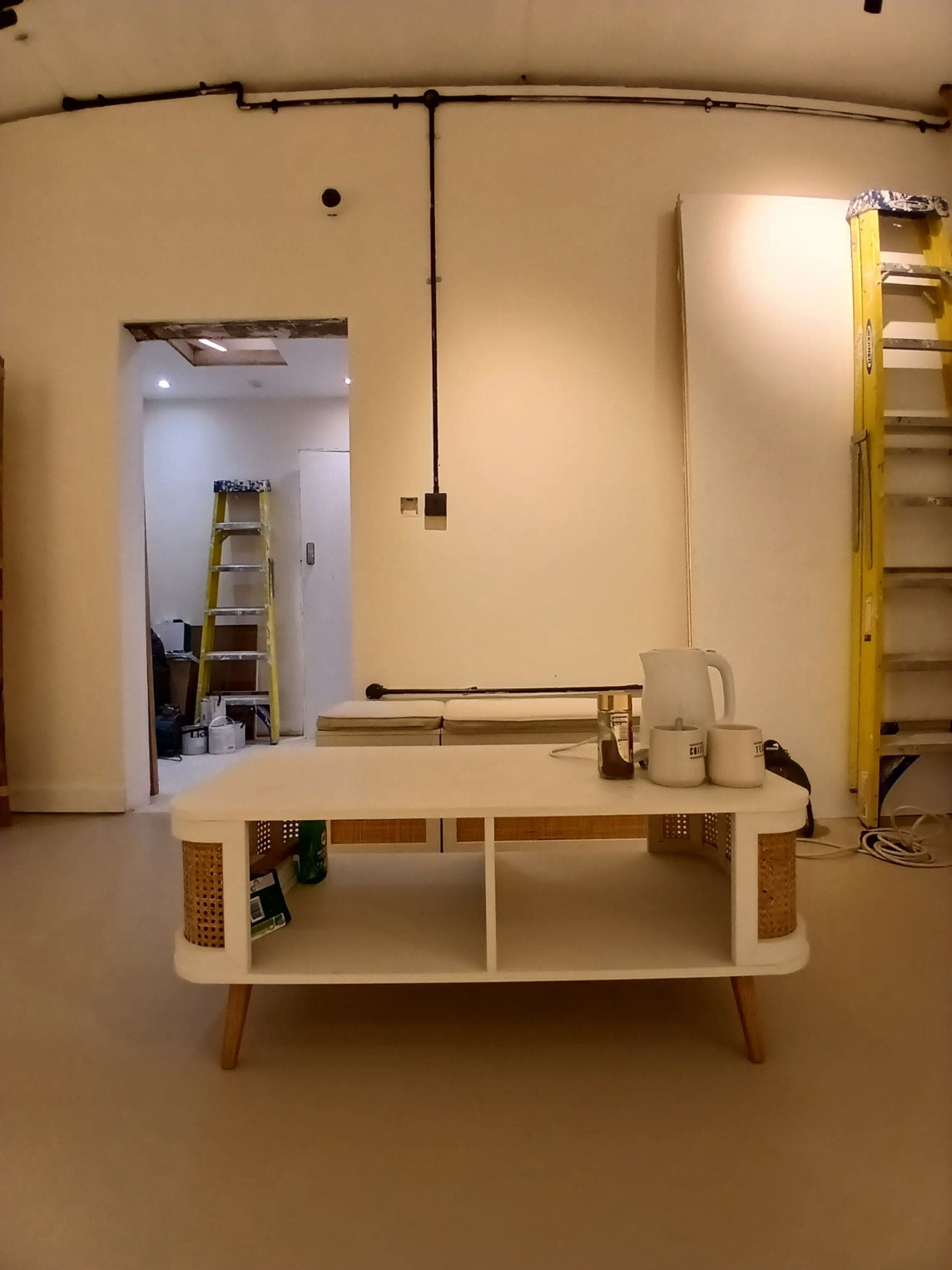 The image shows a minimalistic living space with a coffee table in the foreground, a ladder against the wall, and a partially finished wall in the background.