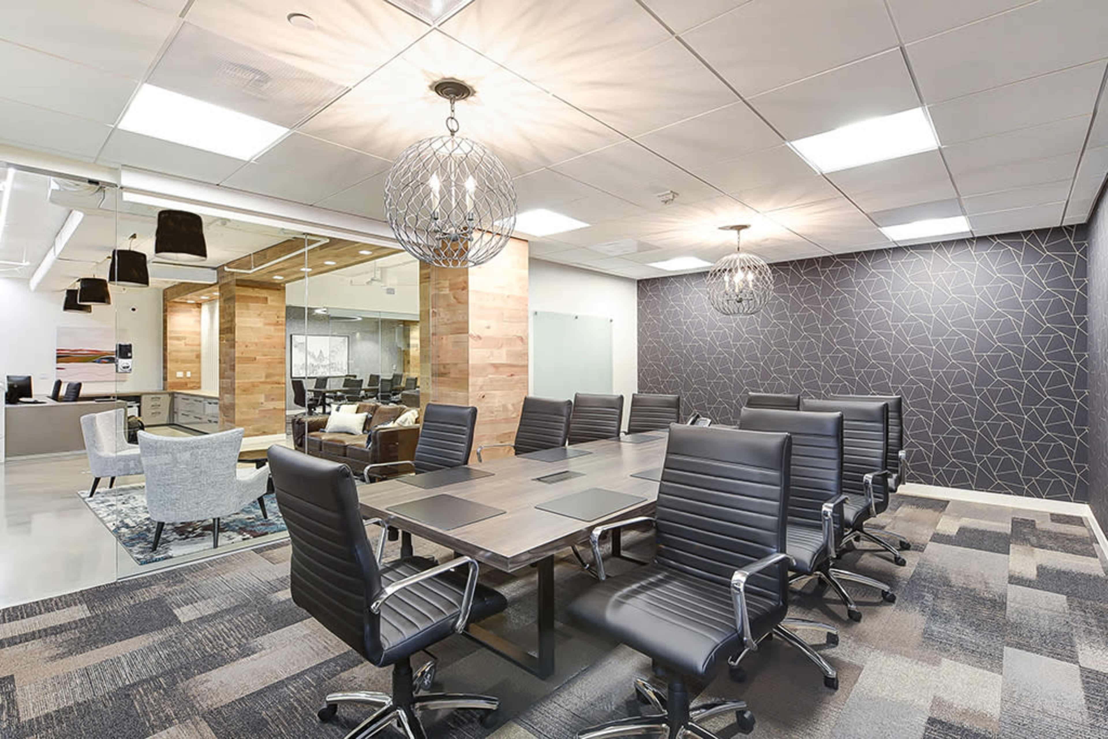 A conference room featuring a long rectangular table surrounded by black office chairs, with decorative light fixtures overhead and a patterned wall.