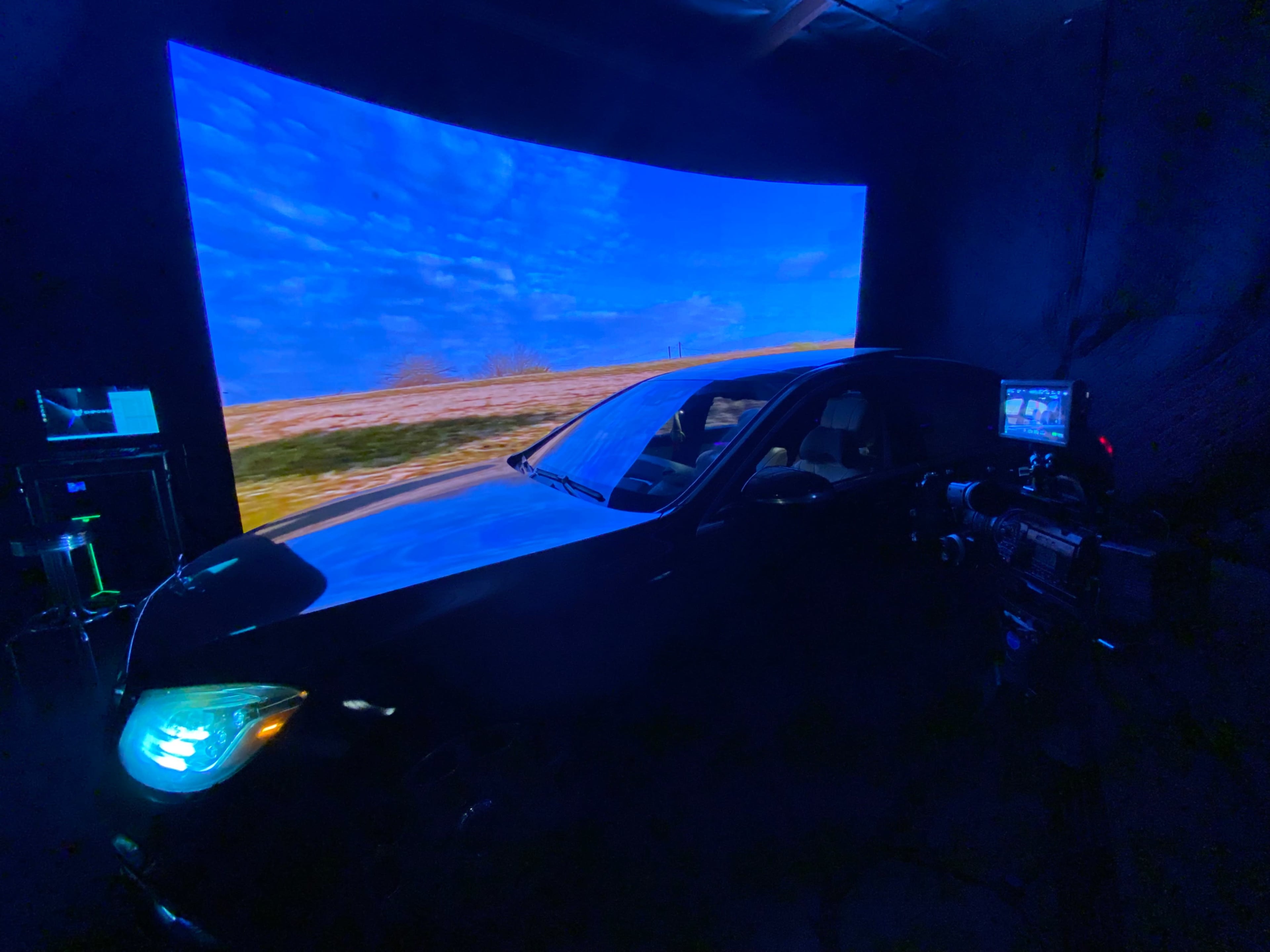 A black car is parked inside a studio with a large curved screen displaying a natural landscape scene.