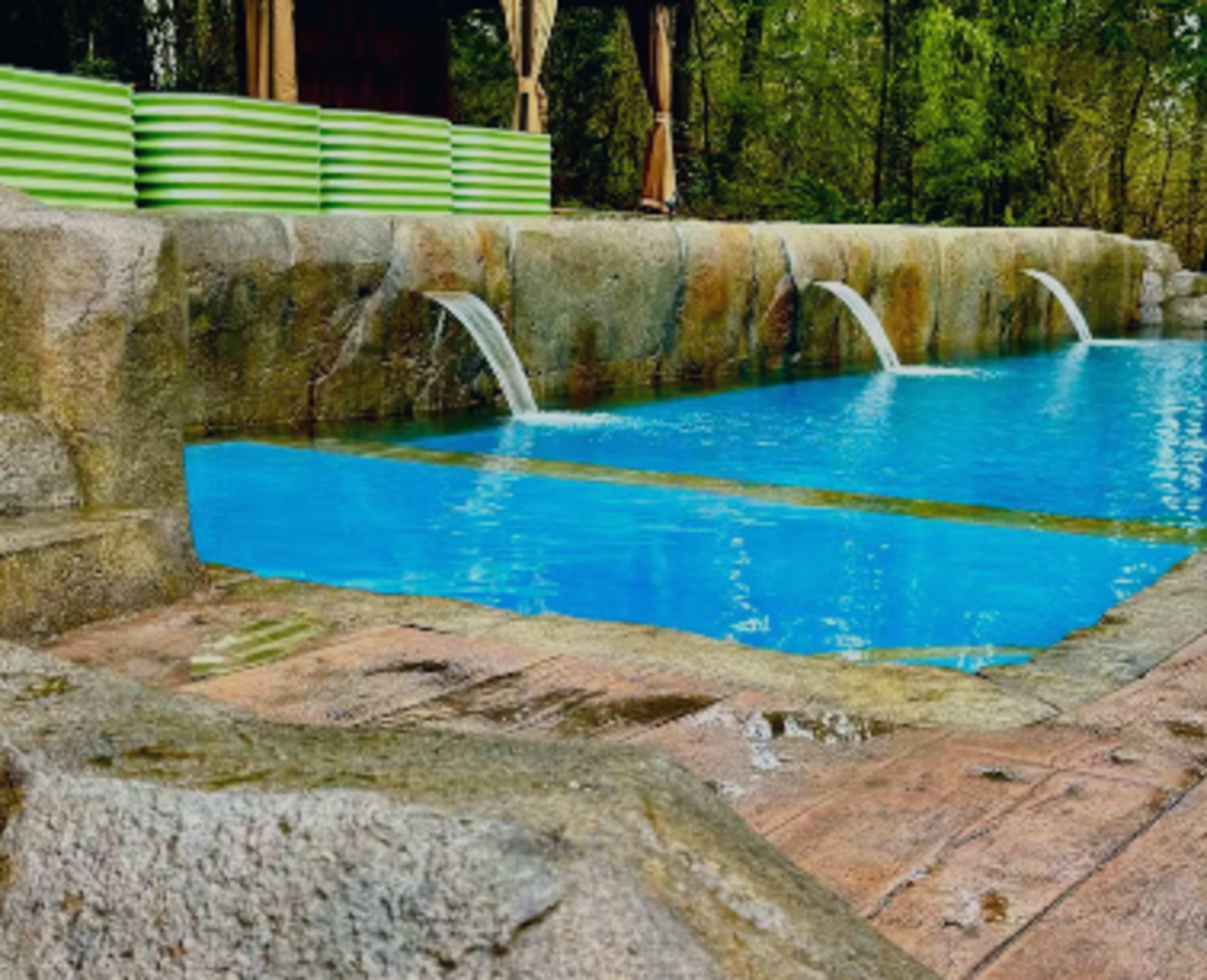 A swimming pool with three water fountains is set against a backdrop of green foliage and wooden decking.