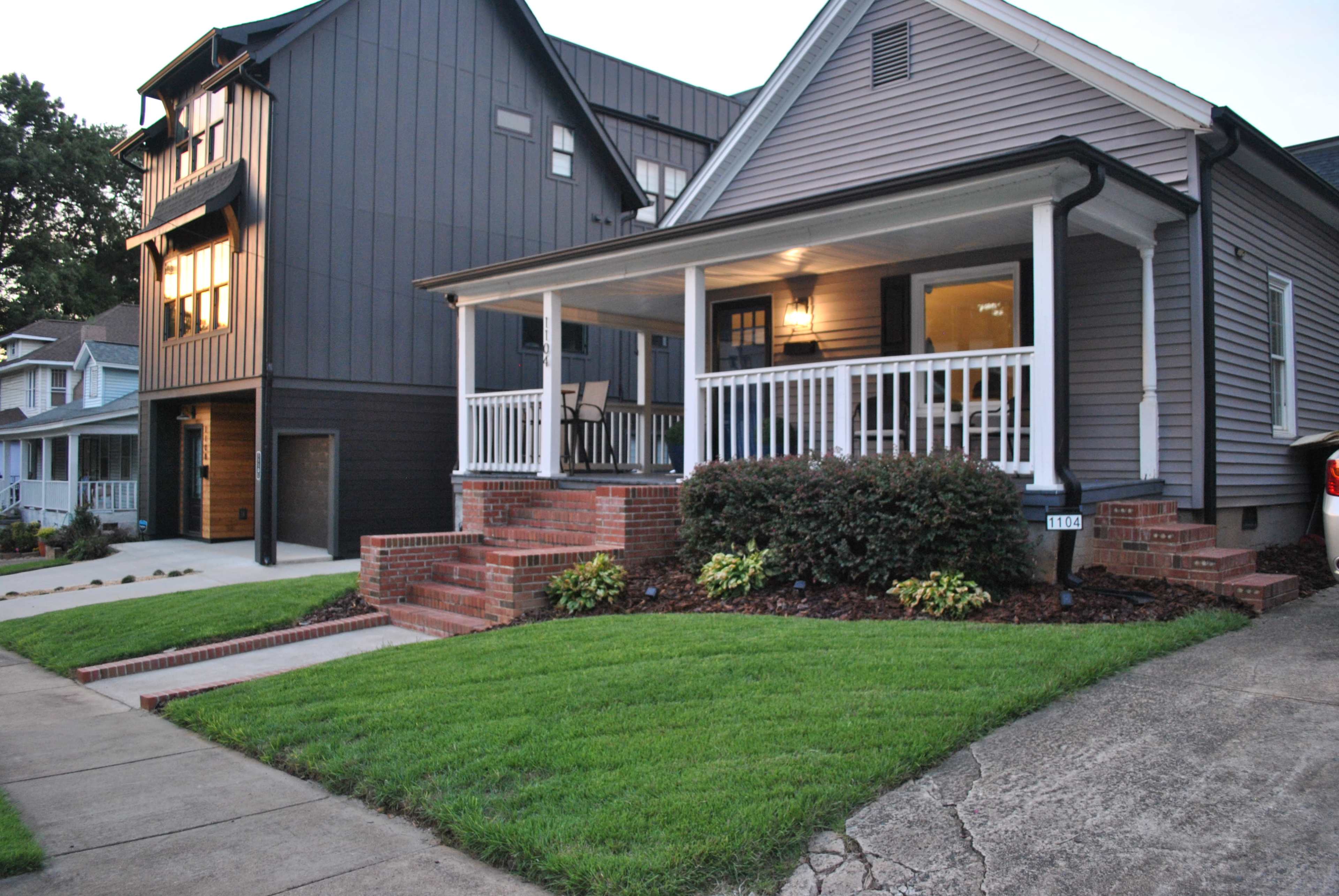 A residential area featuring a gray home with a porch and brick steps, alongside a modern dark-colored house, with well-maintained lawns and sidewalks.