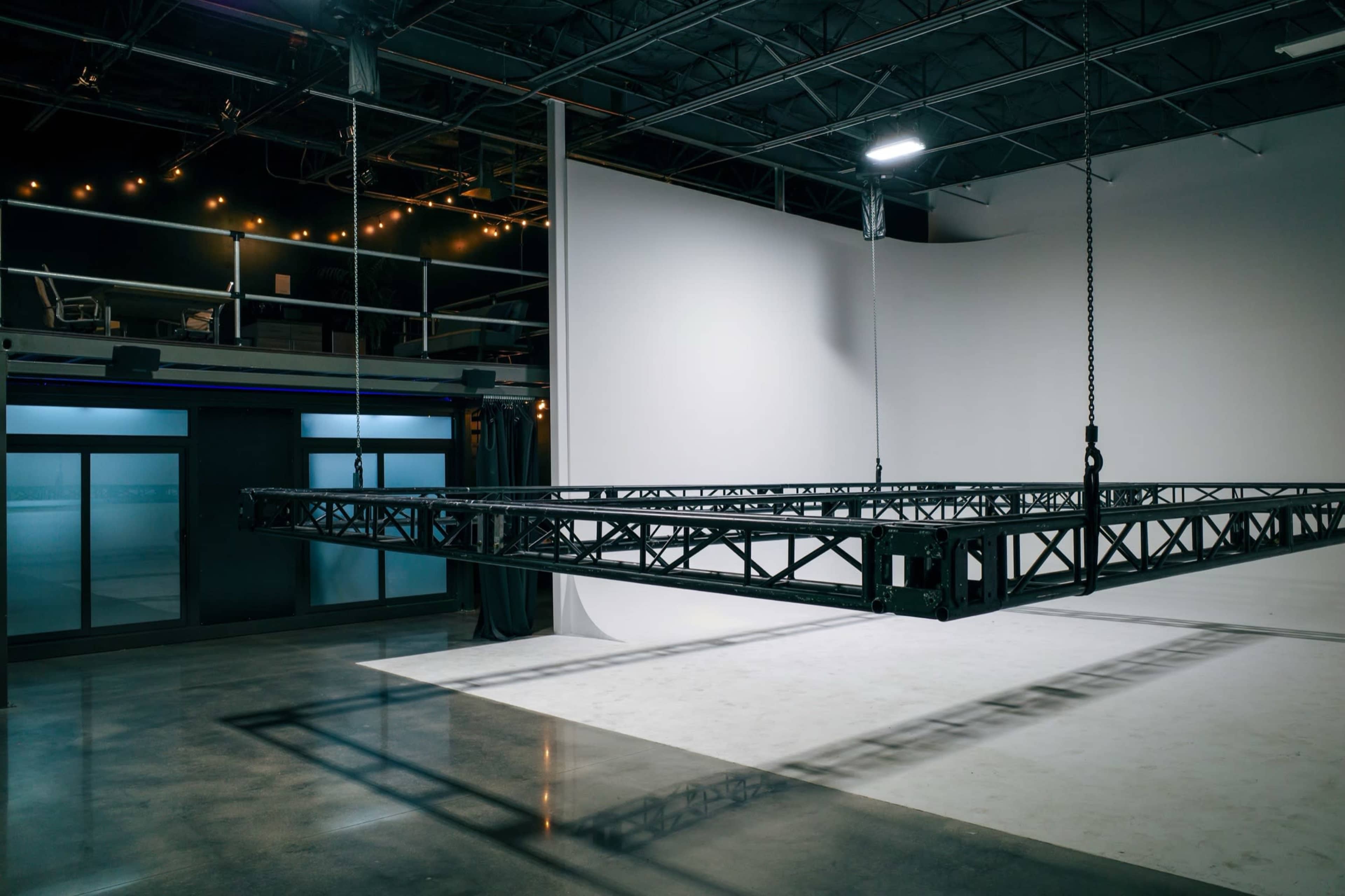 The image shows a large, suspended steel truss framework in an empty studio space with a white backdrop.
