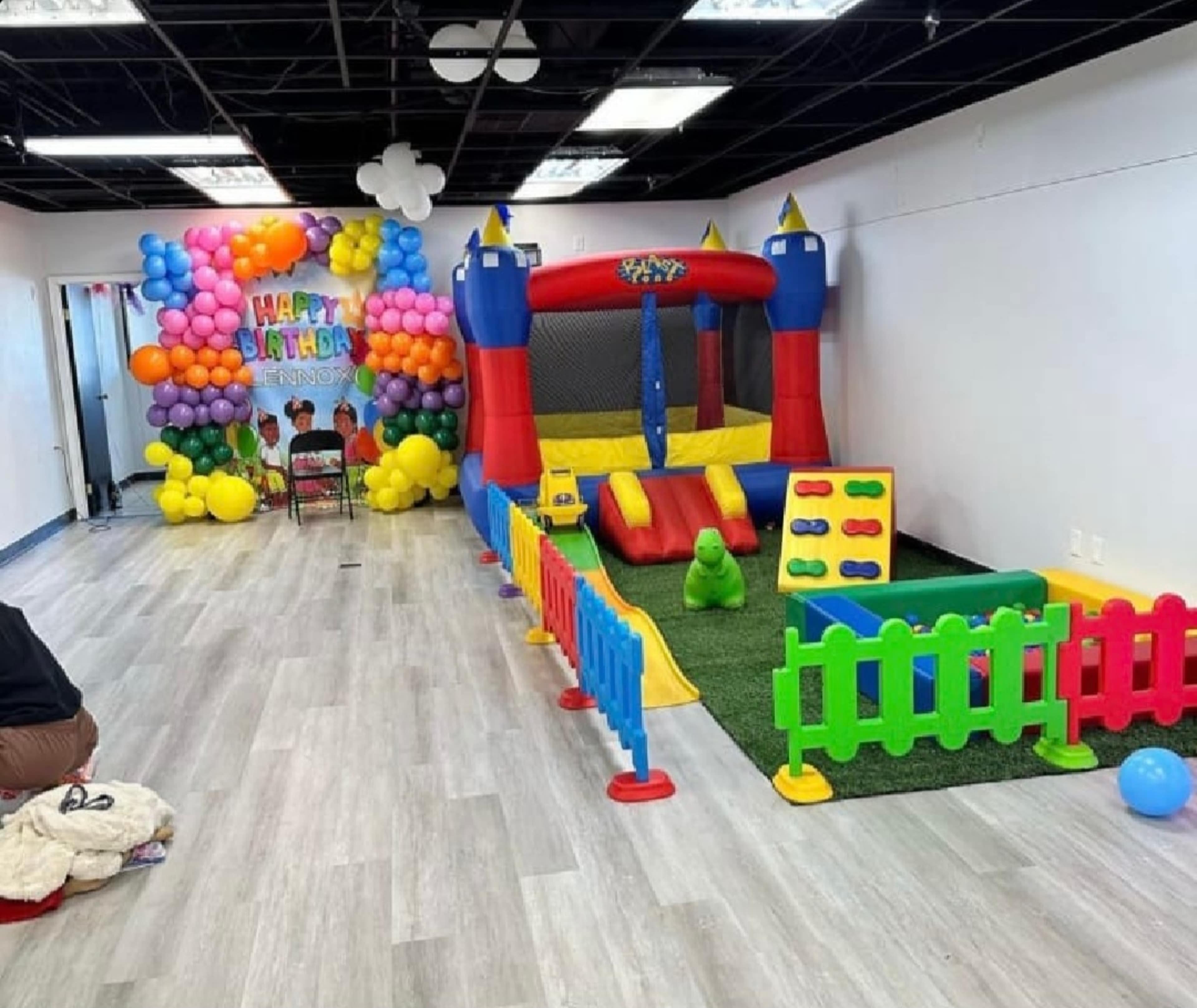 A colorful indoor party area with a bouncy castle, a play area surrounded by a toy fence, and a balloon backdrop that reads "Happy Birthday."