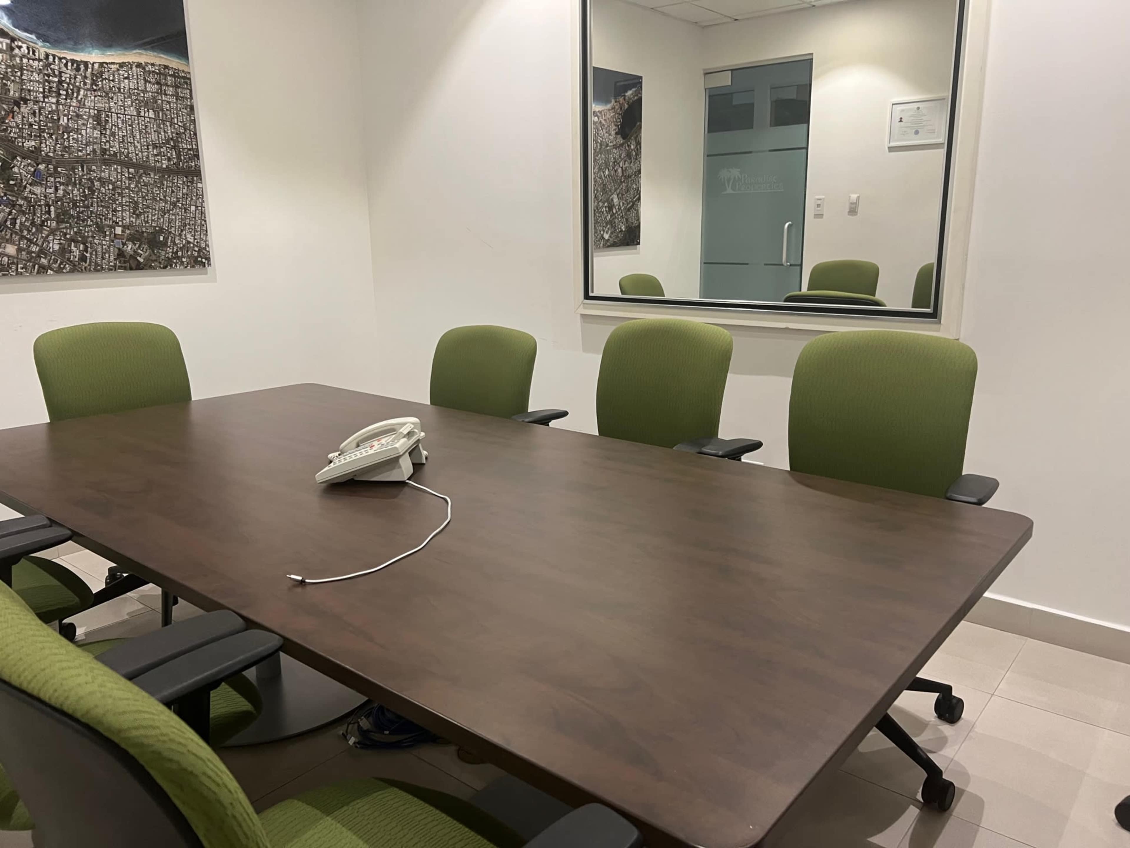 A conference room features a rectangular brown table with five green chairs and a white telephone on the surface.