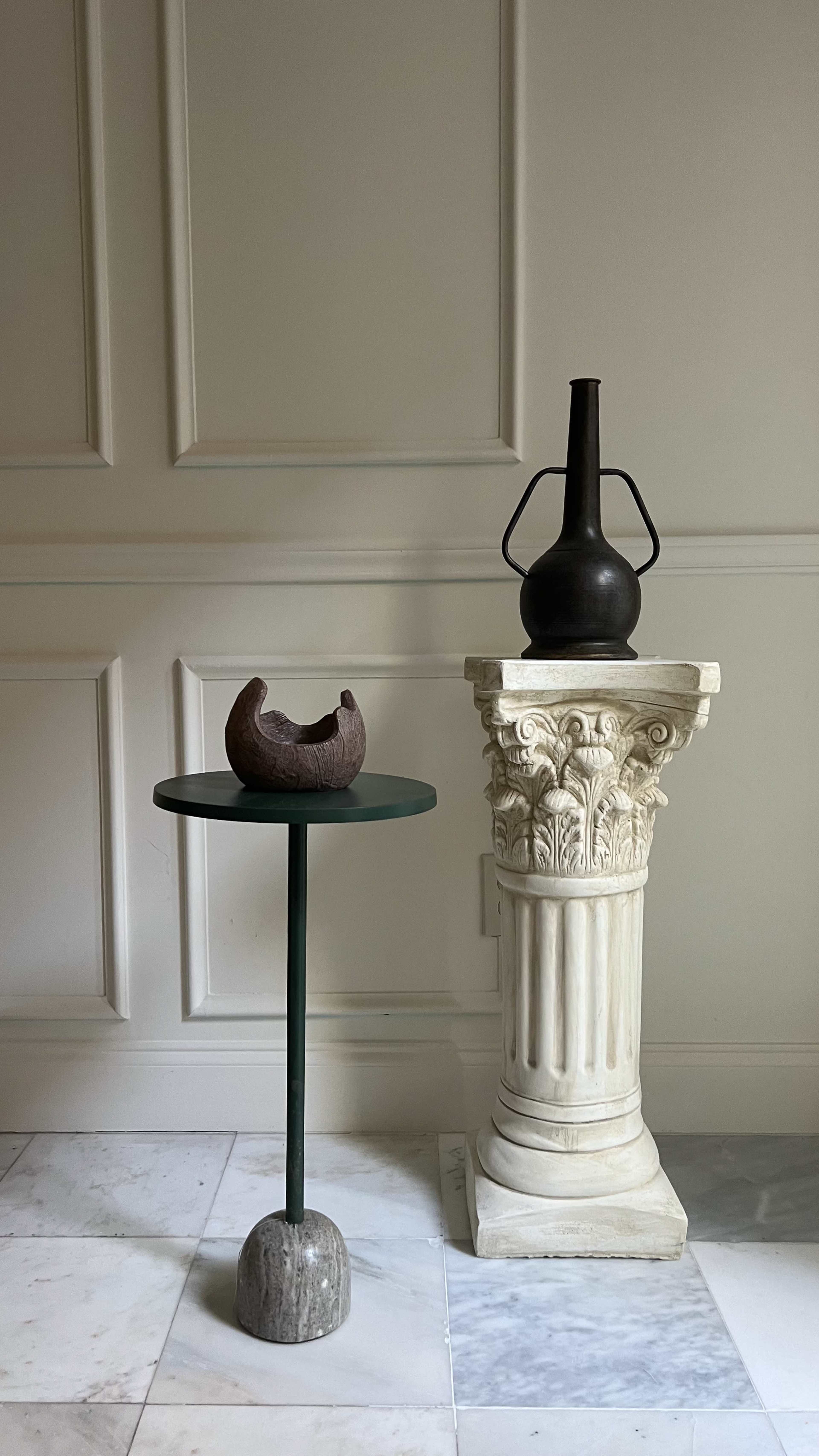 A minimalist interior scene featuring a black vase on a classical column beside a round green table with a textured, dark bowl resting on it.