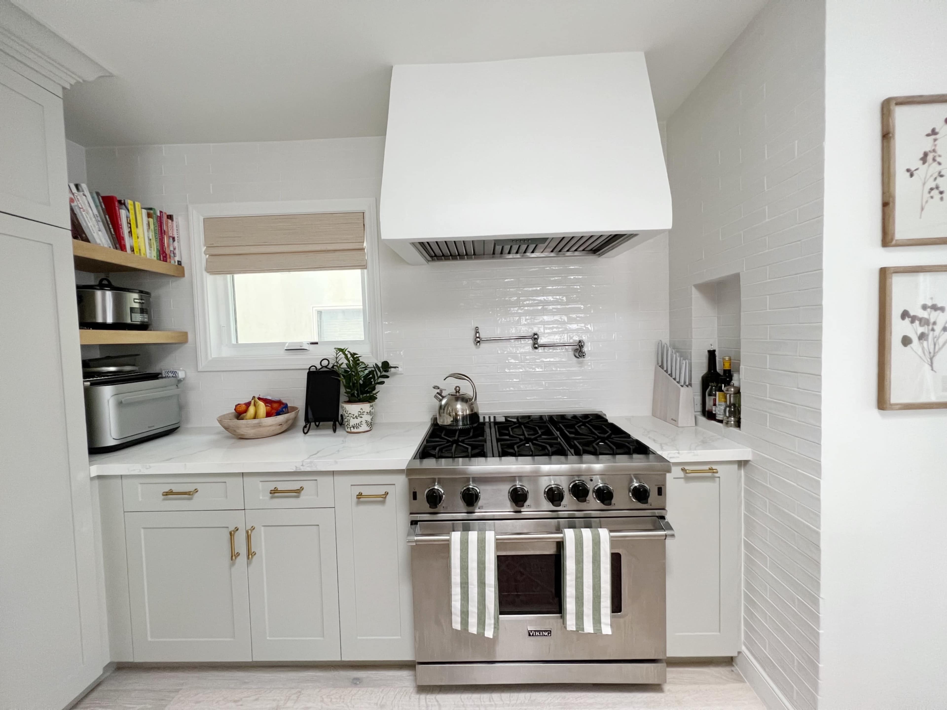 A modern kitchen featuring a professional-grade gas stove, white cabinetry, open shelving with cookbooks, and a stainless steel range hood.