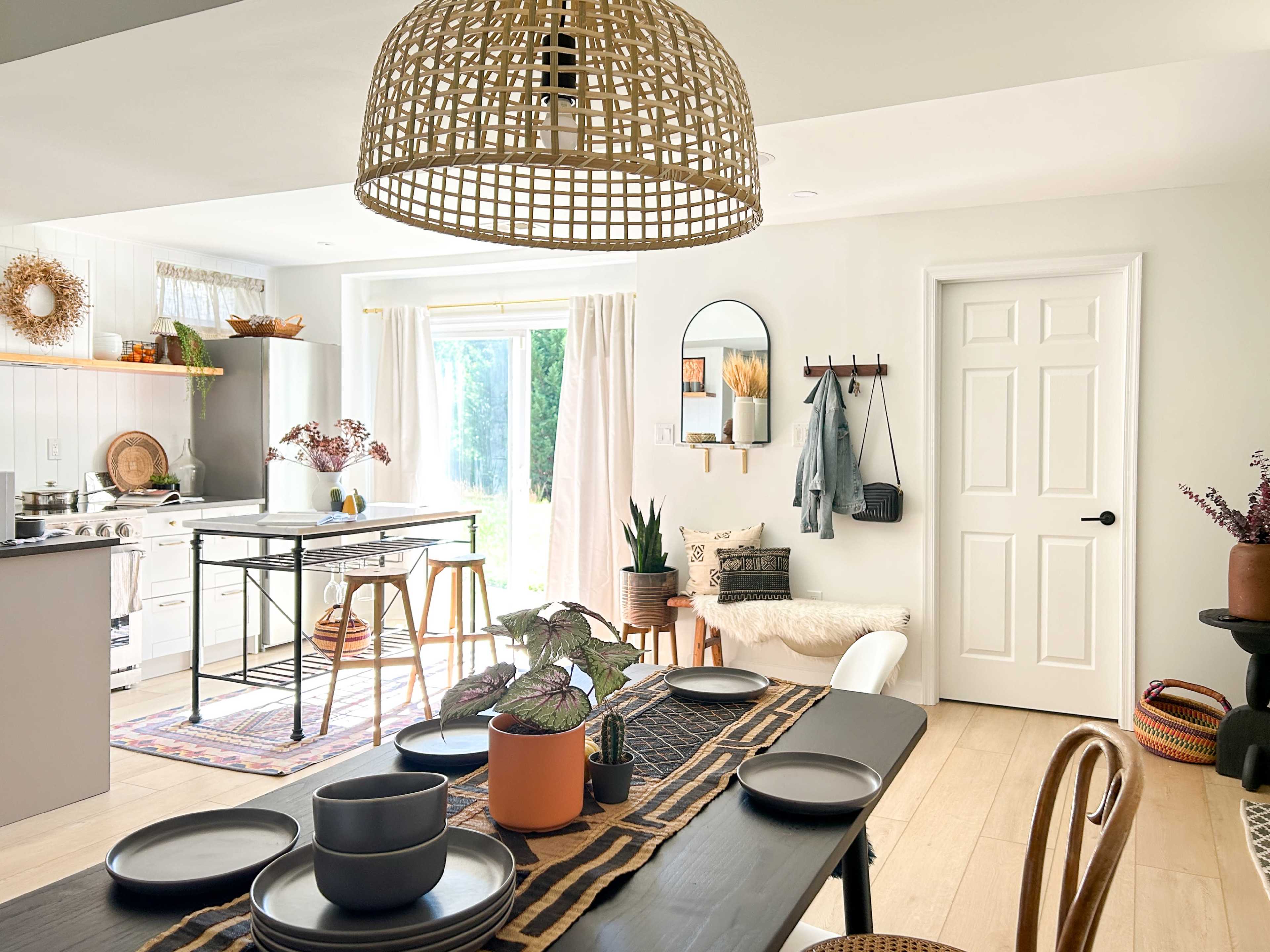 A bright dining area features a long black table set with plates, surrounded by stylish seating and a view of greenery through large glass doors.