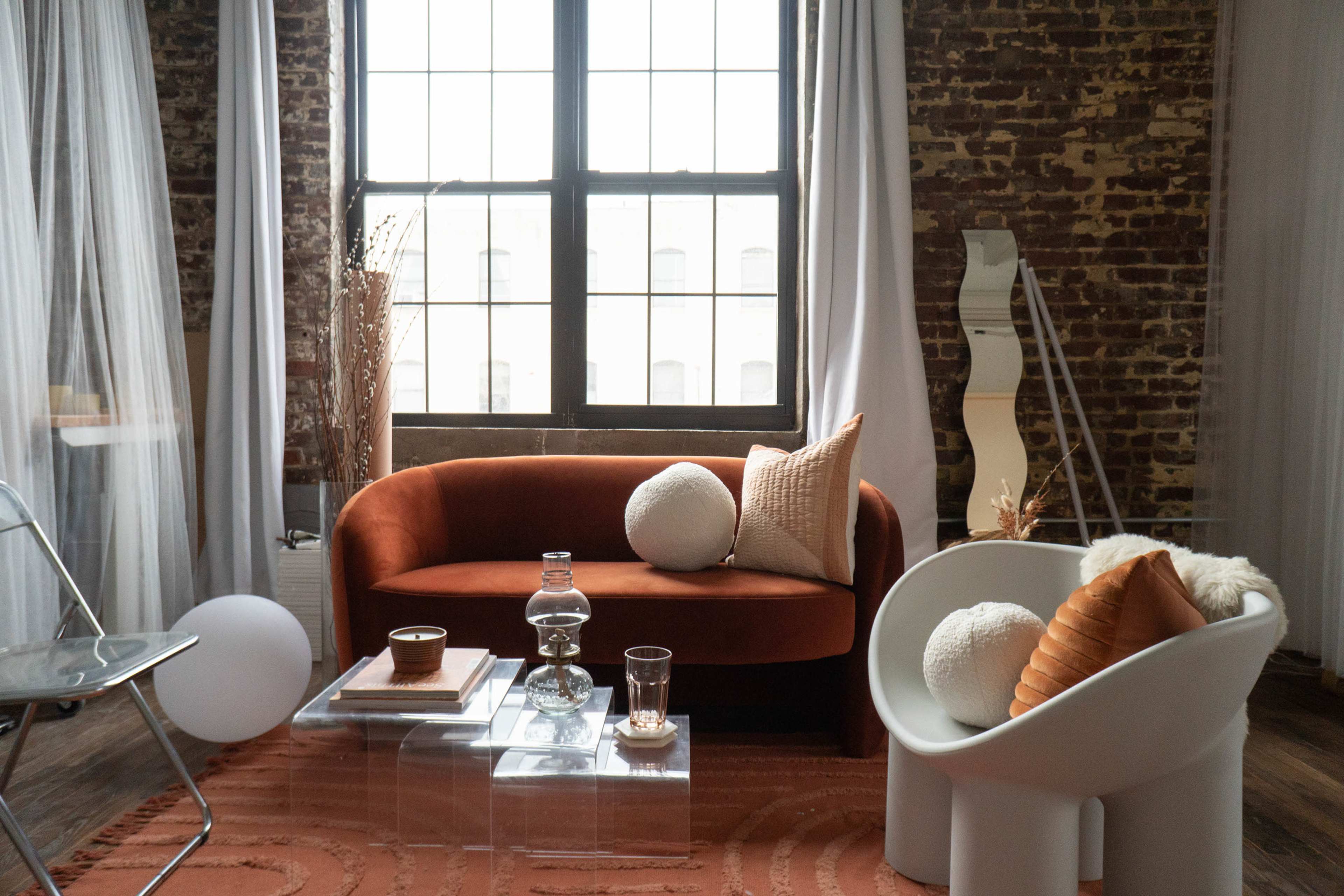 A cozy living space with a brick wall, featuring an orange sofa, a white sculptural chair, and a glass coffee table with decorative items.