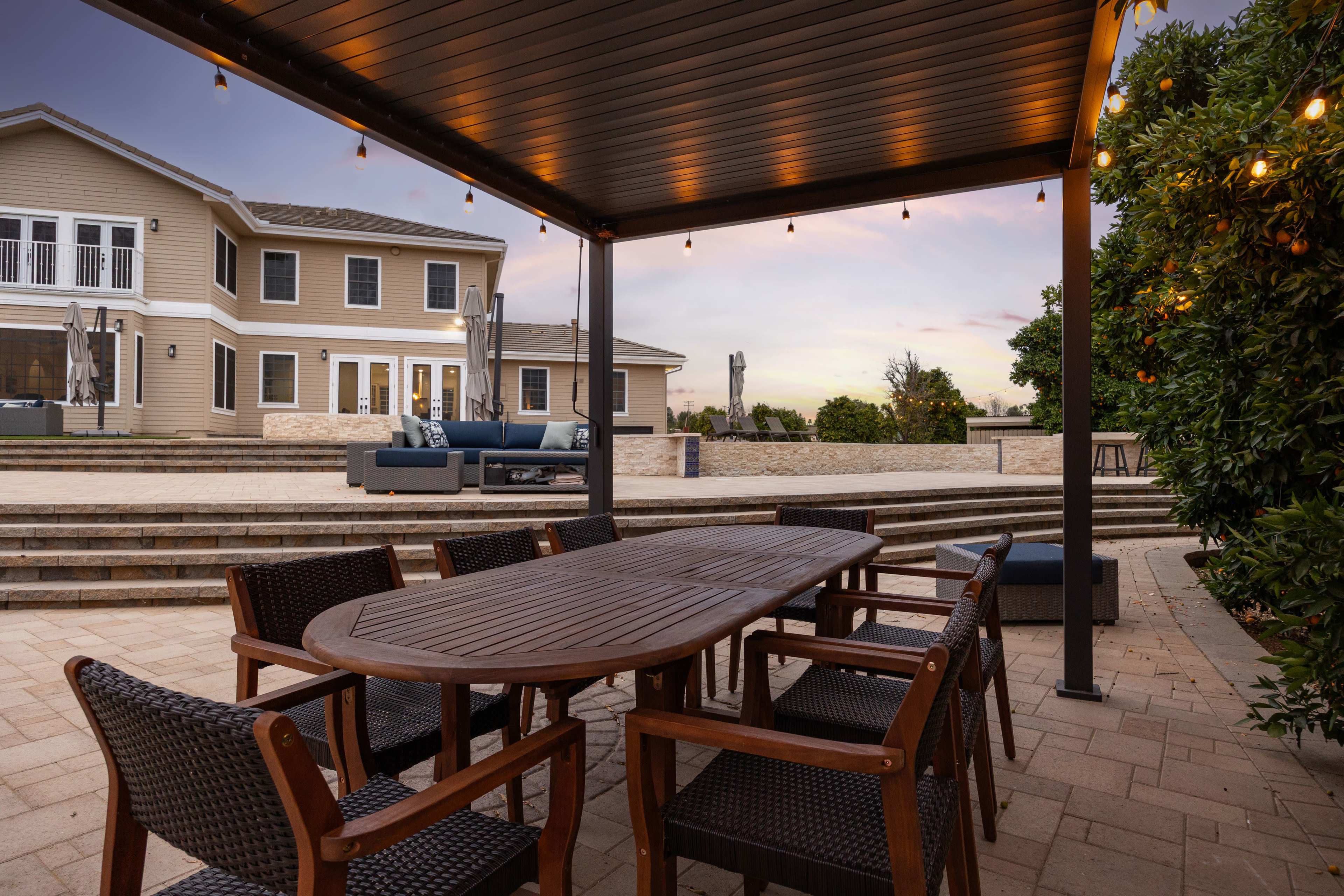 A wooden dining table with chairs is set under a pergola, overlooking a landscaped backyard and a multi-story house.