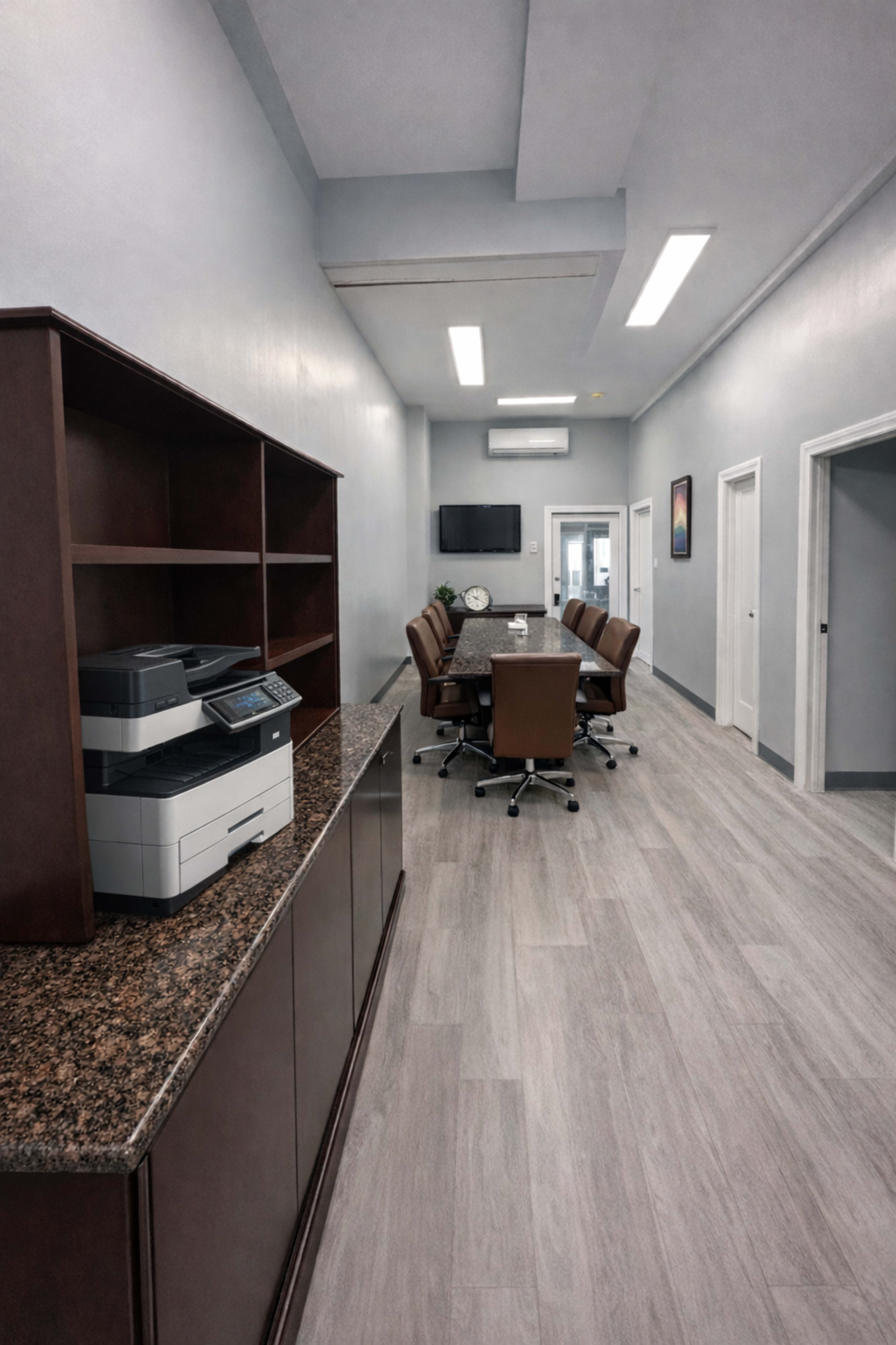 The image shows a long, narrow office space featuring a conference table with chairs, a printer on a cabinet, and a television mounted on the wall.