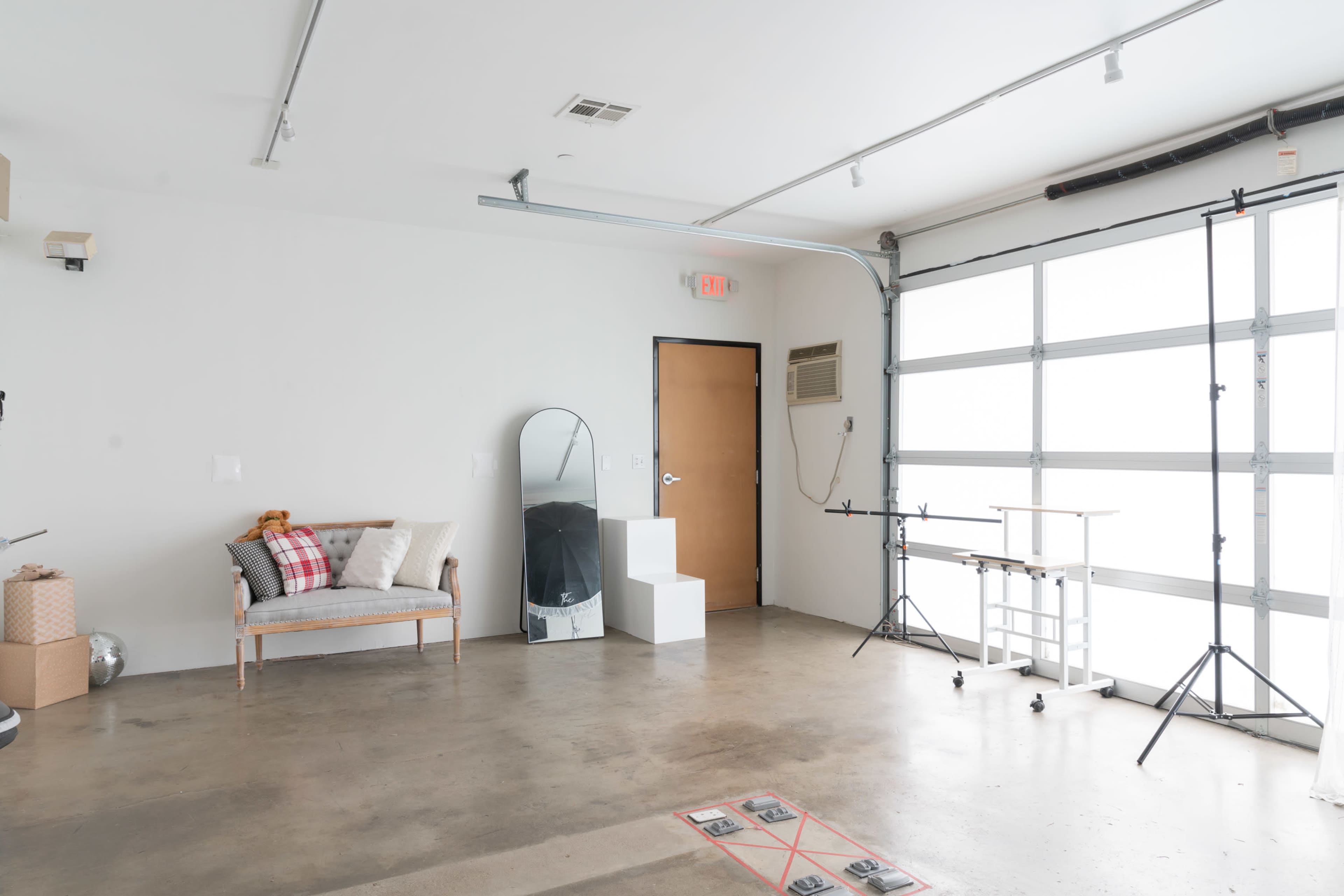An empty room with a polished concrete floor, featuring a sofa, a large mirror, a white pedestal, and equipment for photography along one wall.