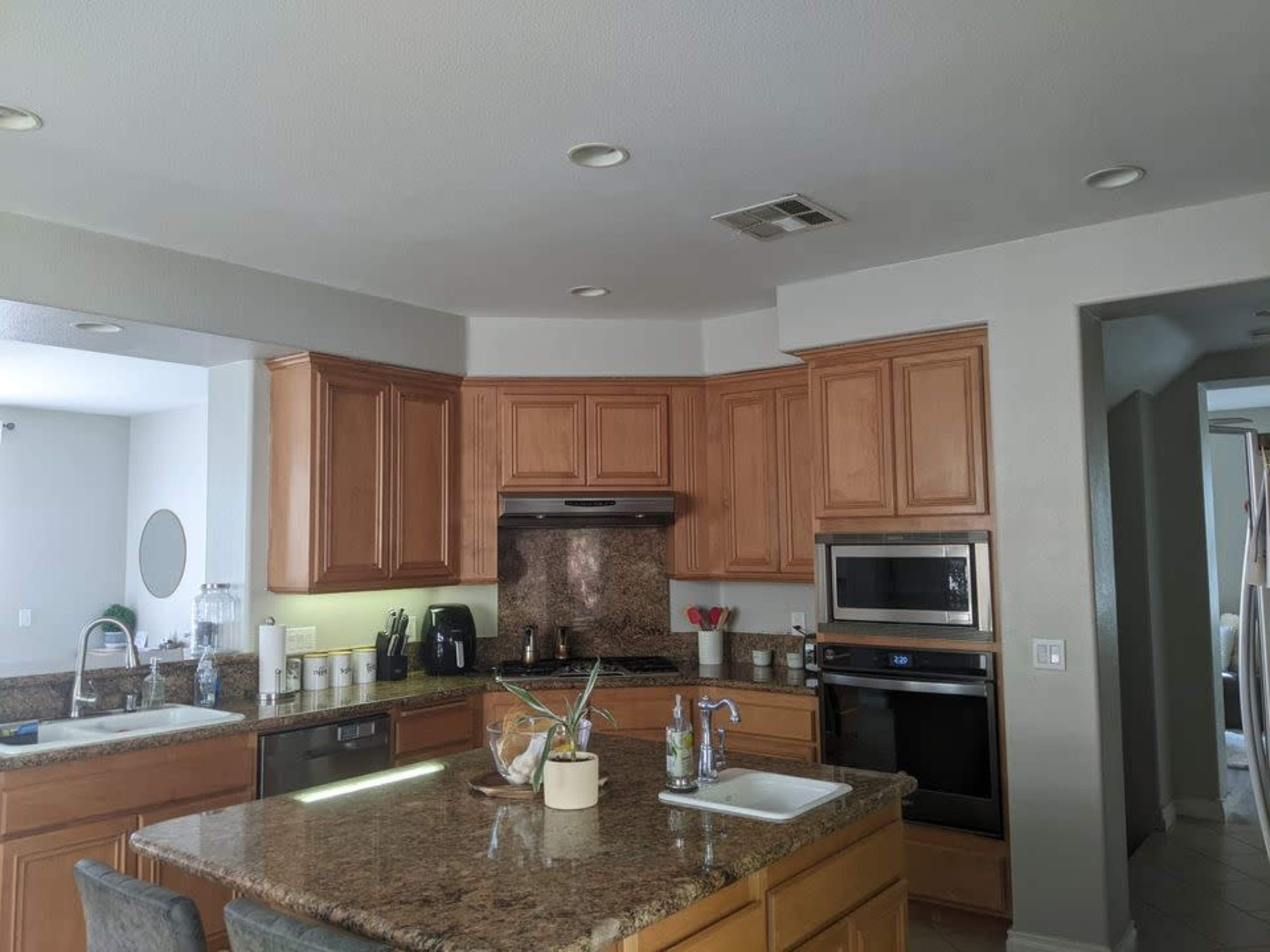 The image shows a kitchen with wooden cabinetry, granite countertops, modern appliances, and an island with seating.