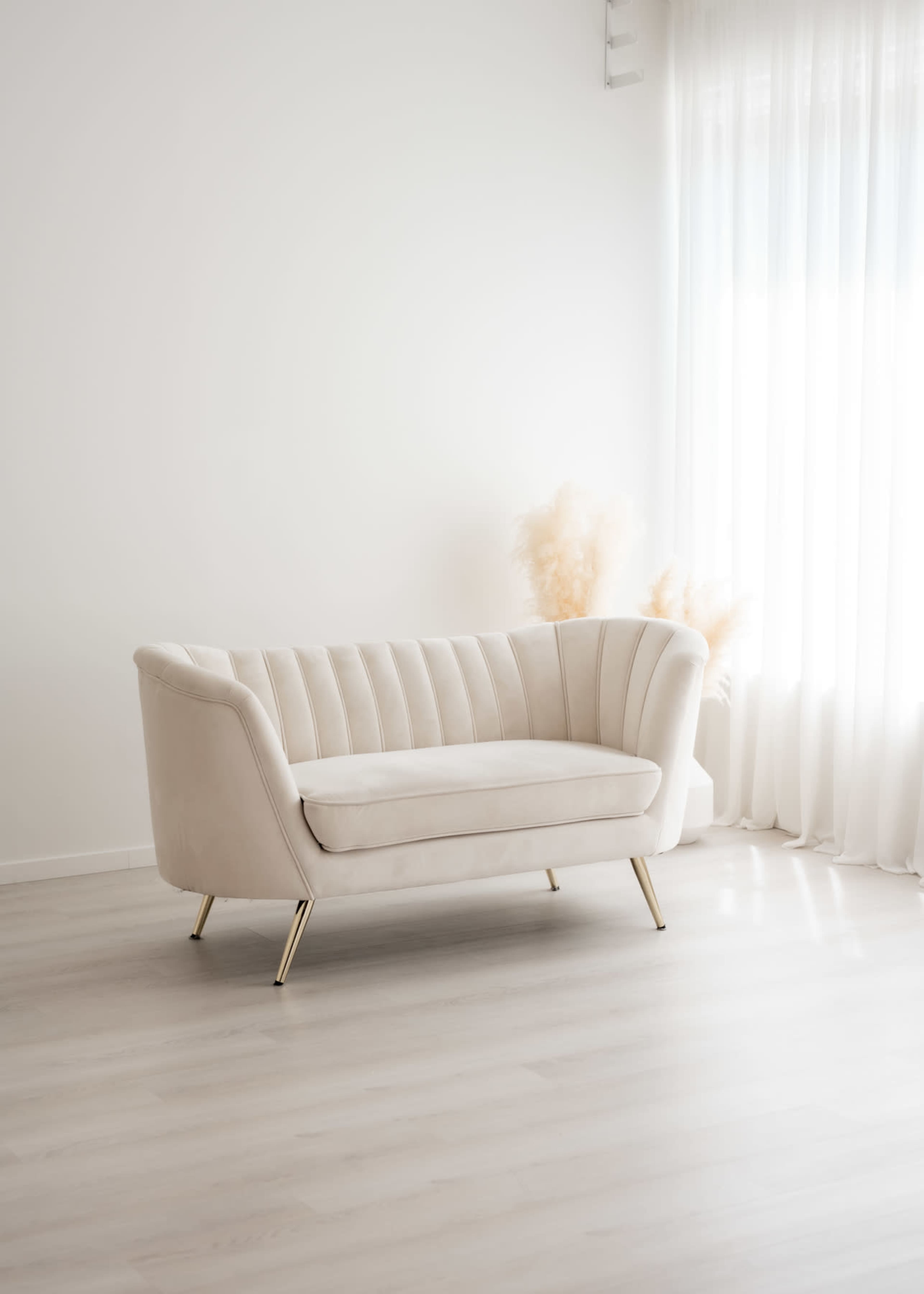 A light-colored, modern couch with a curved design and gold legs is positioned in a bright, minimalist room next to a window with sheer curtains.