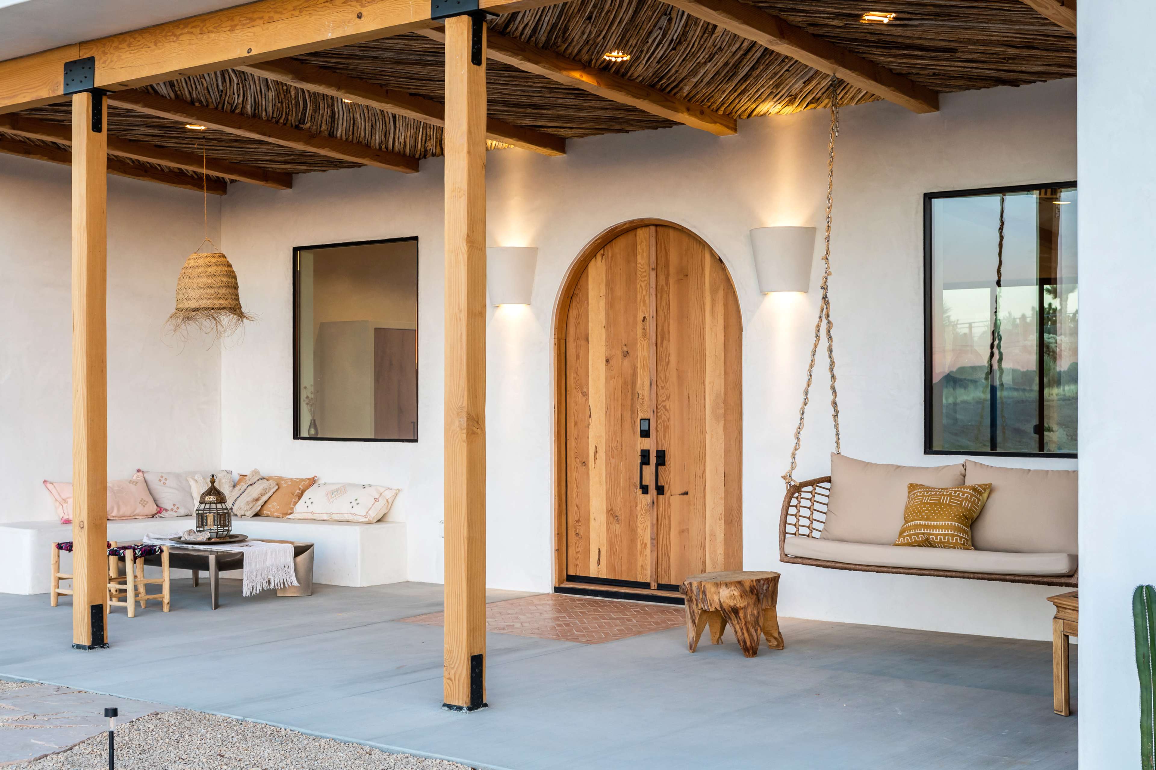 The image shows a modern outdoor porch featuring a wooden front door, a swing seat, and a seating area with decorative pillows beneath a thatched roof.