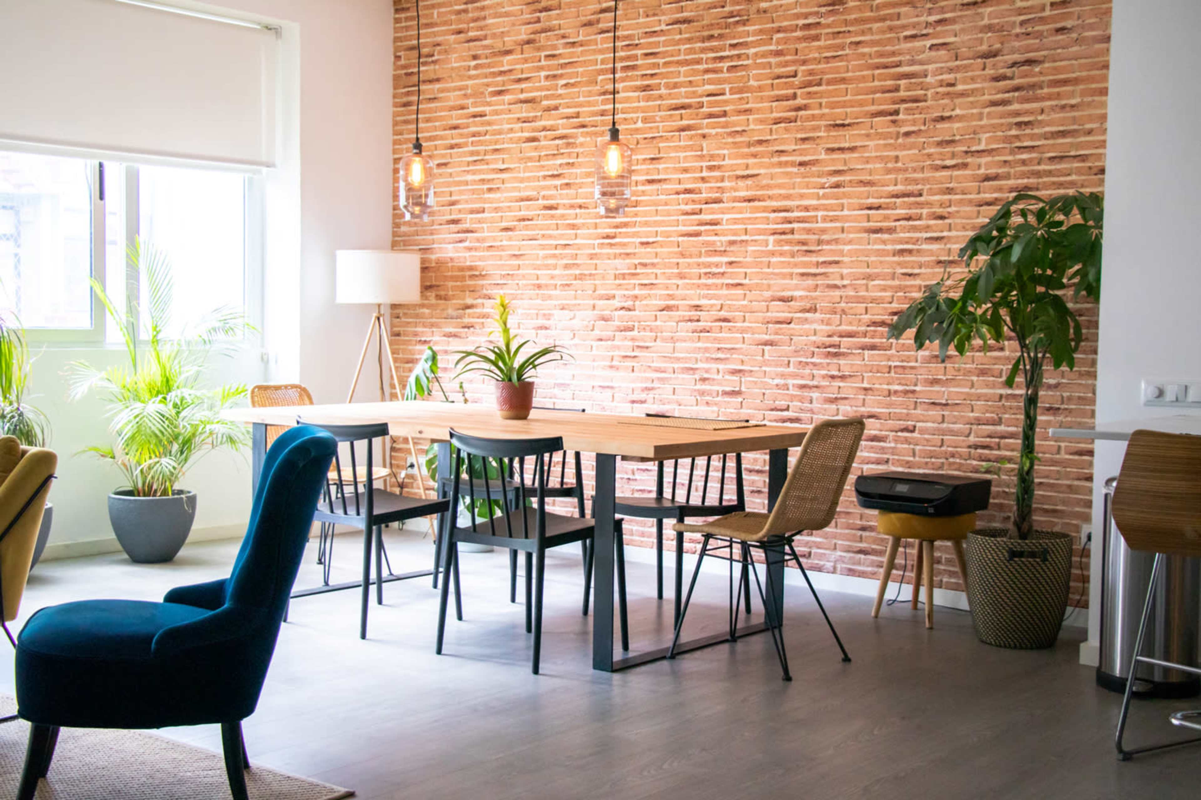 The image shows a modern dining space with a large wooden table surrounded by chairs, a brick wall in the background, and potted plants in the corners.