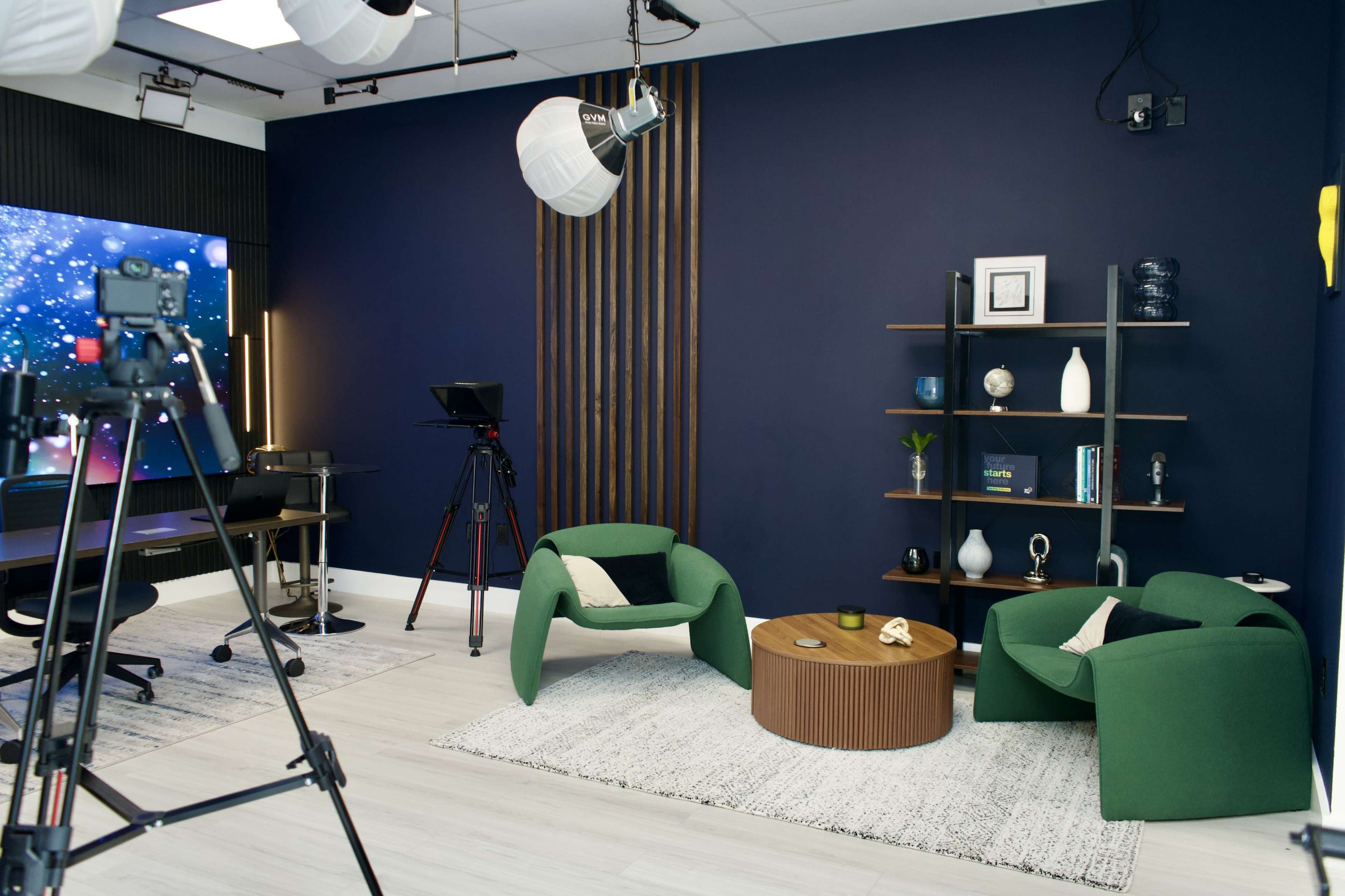 The image shows a modern studio setup featuring two green chairs, a round wooden table, a shelving unit with decorative items, and multiple cameras and lighting equipment.