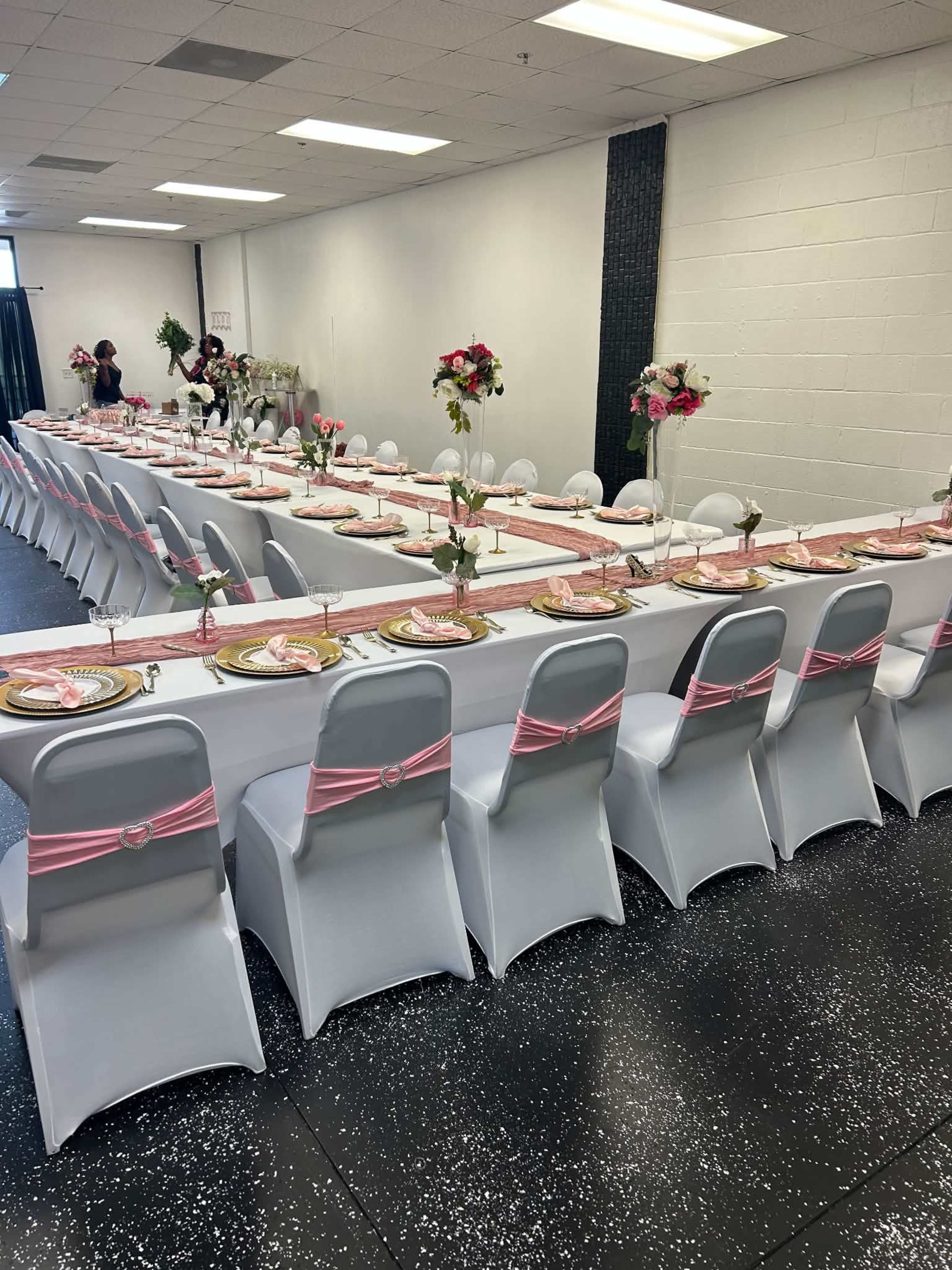 A long table is elegantly set with pink and gold decorations, surrounded by white chairs, in a decorated indoor venue.