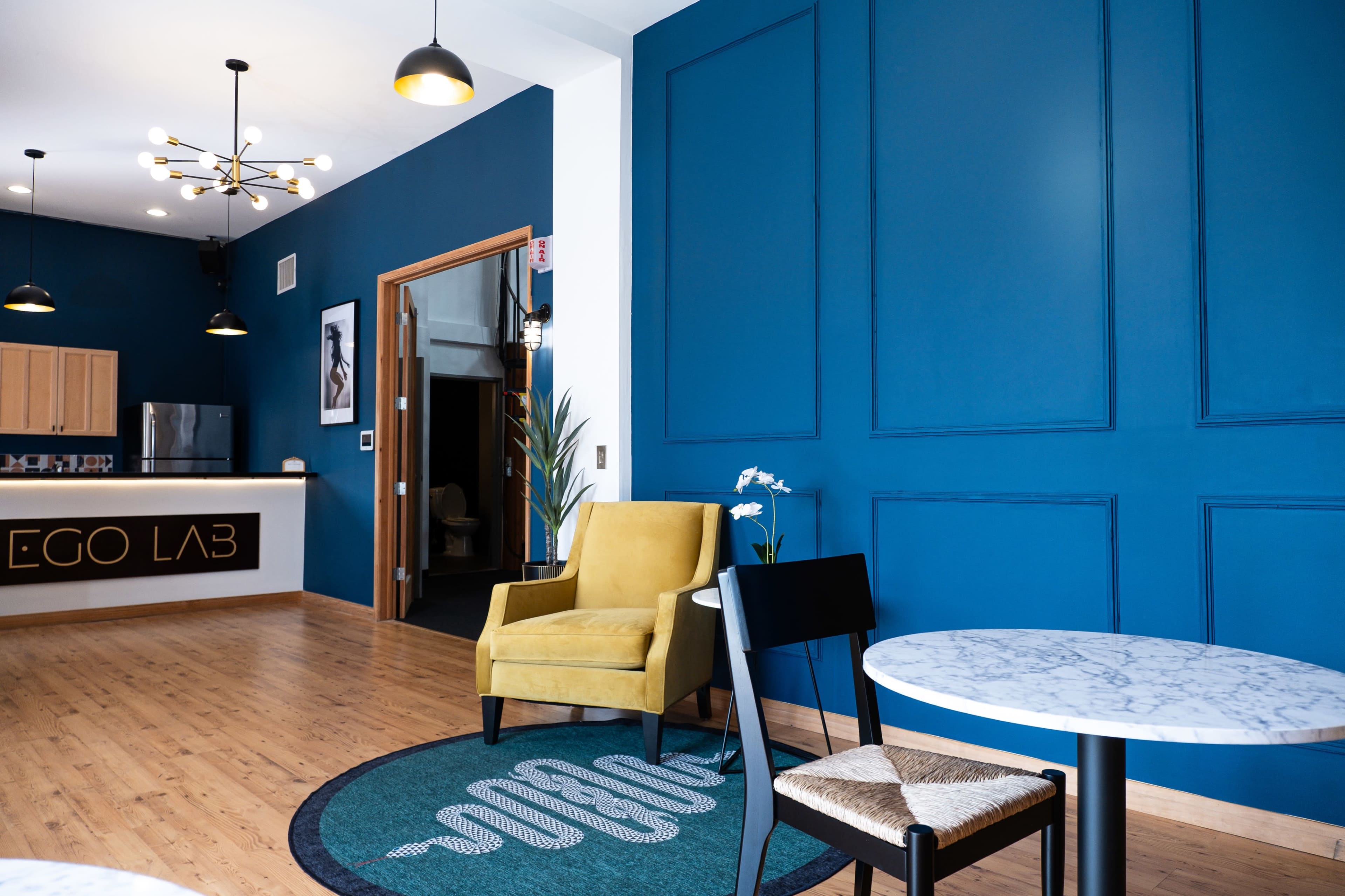 A modern interior featuring a blue wall, a yellow armchair, a marble-topped table, and a reception area with a sign that reads "EGO LAB."