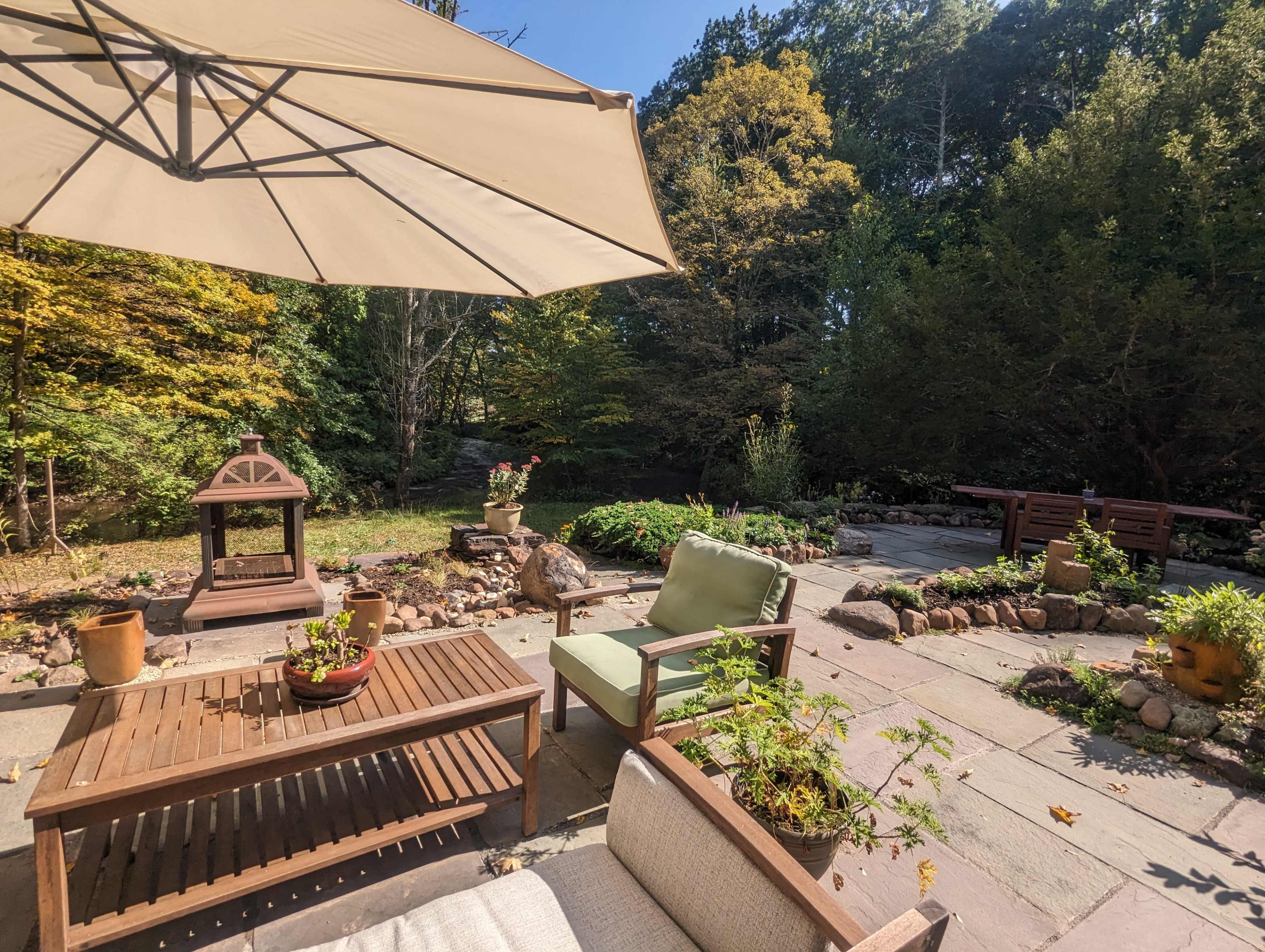 A patio area features wooden furniture, a large umbrella, and surrounding lush greenery.