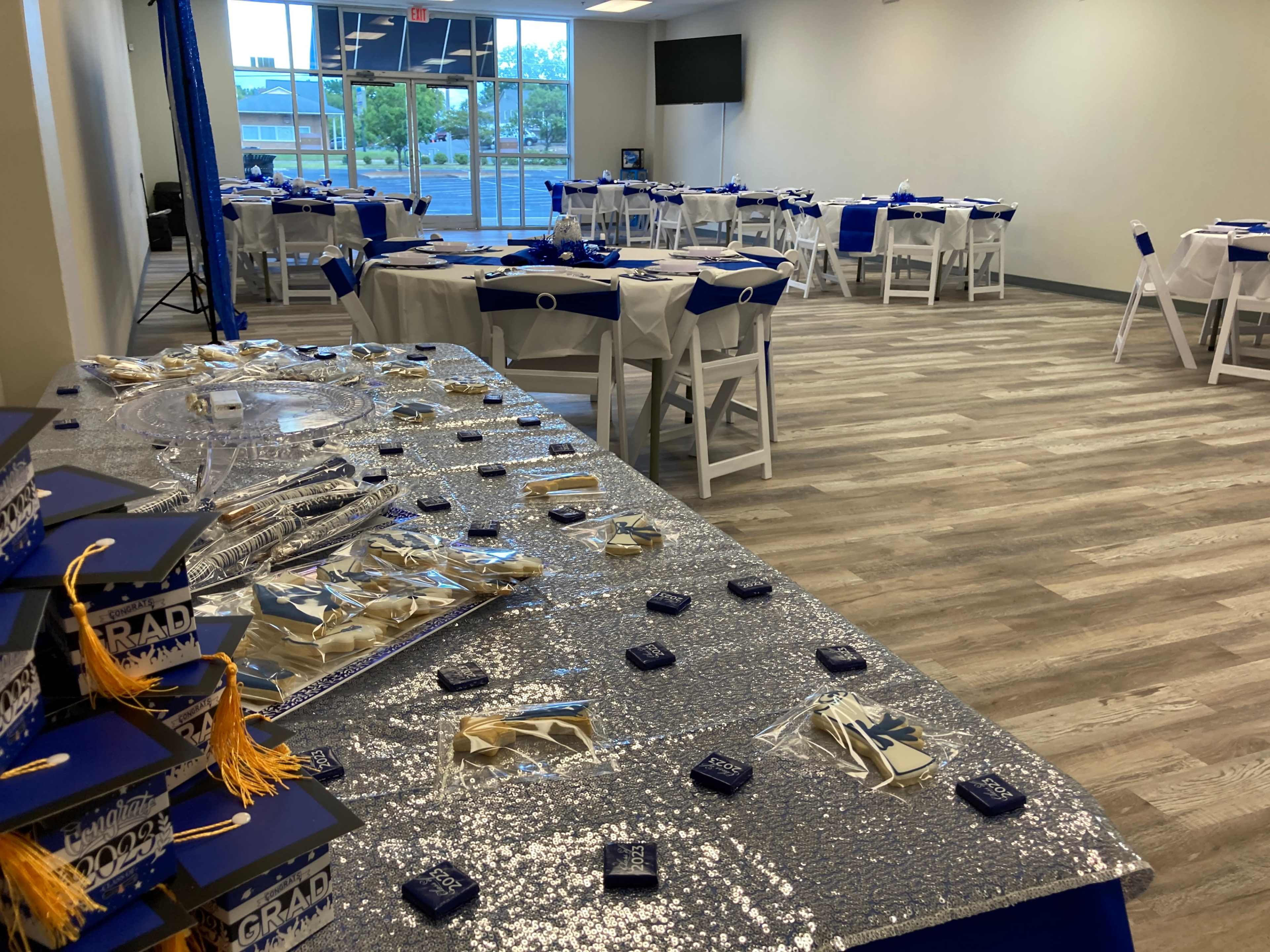 The image shows a decorated event space with tables set for a graduation celebration, featuring blue and silver decor and a display of graduation caps.