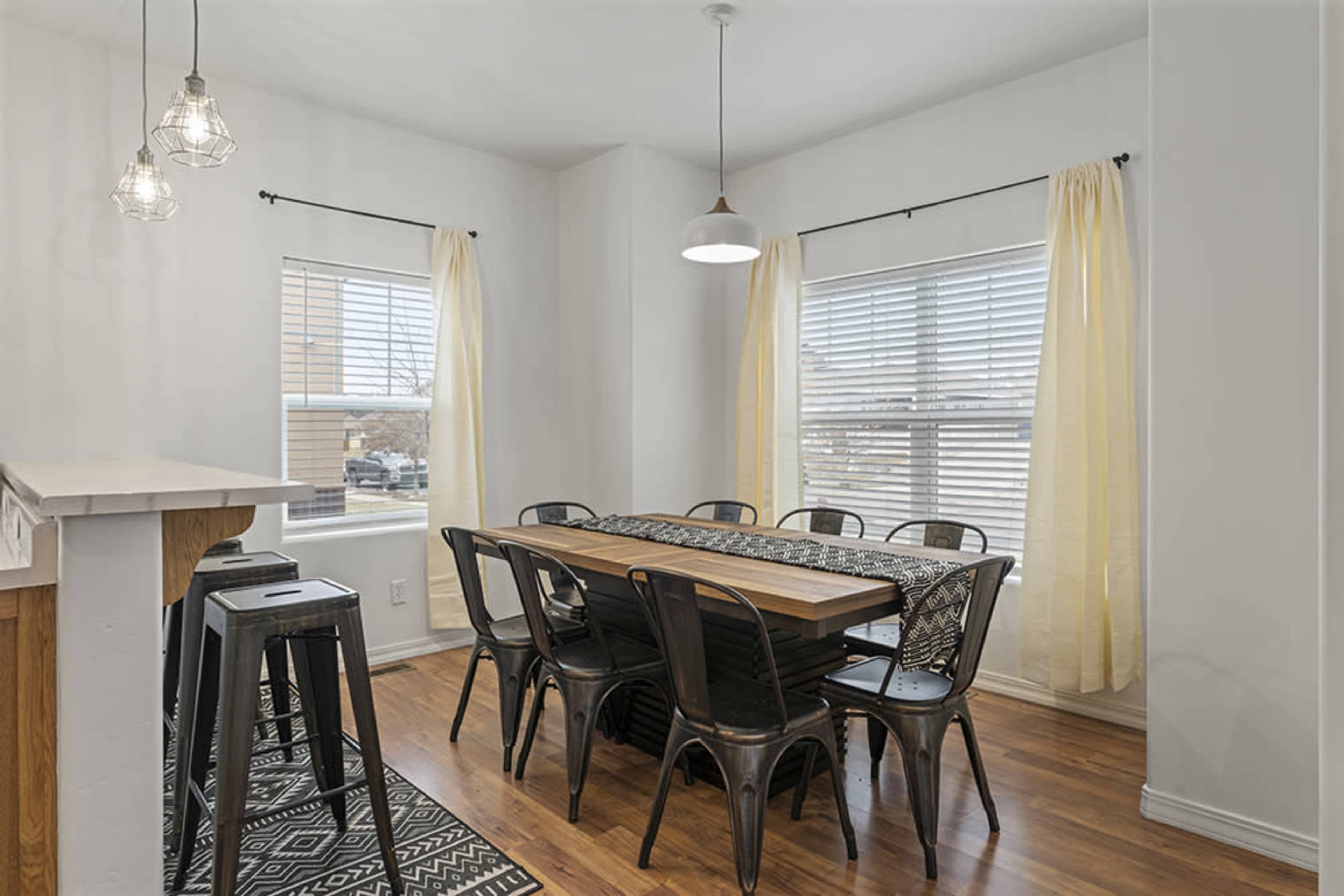A dining area features a long wooden table surrounded by metal chairs, illuminated by pendant lights and natural light from two windows.
