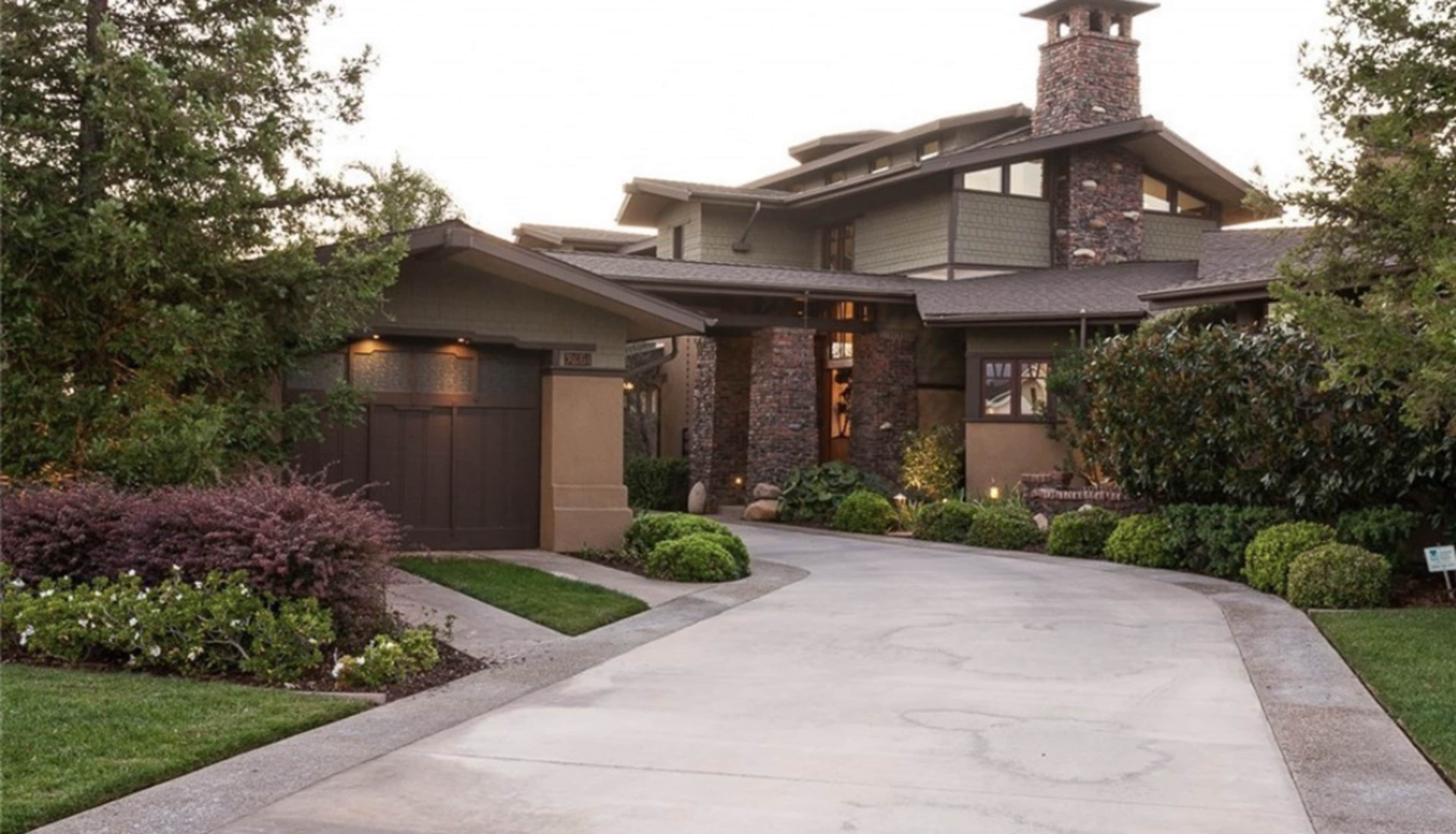 The image shows a modern two-story house with stone and wood exterior, featuring a large driveway and landscaped greenery.